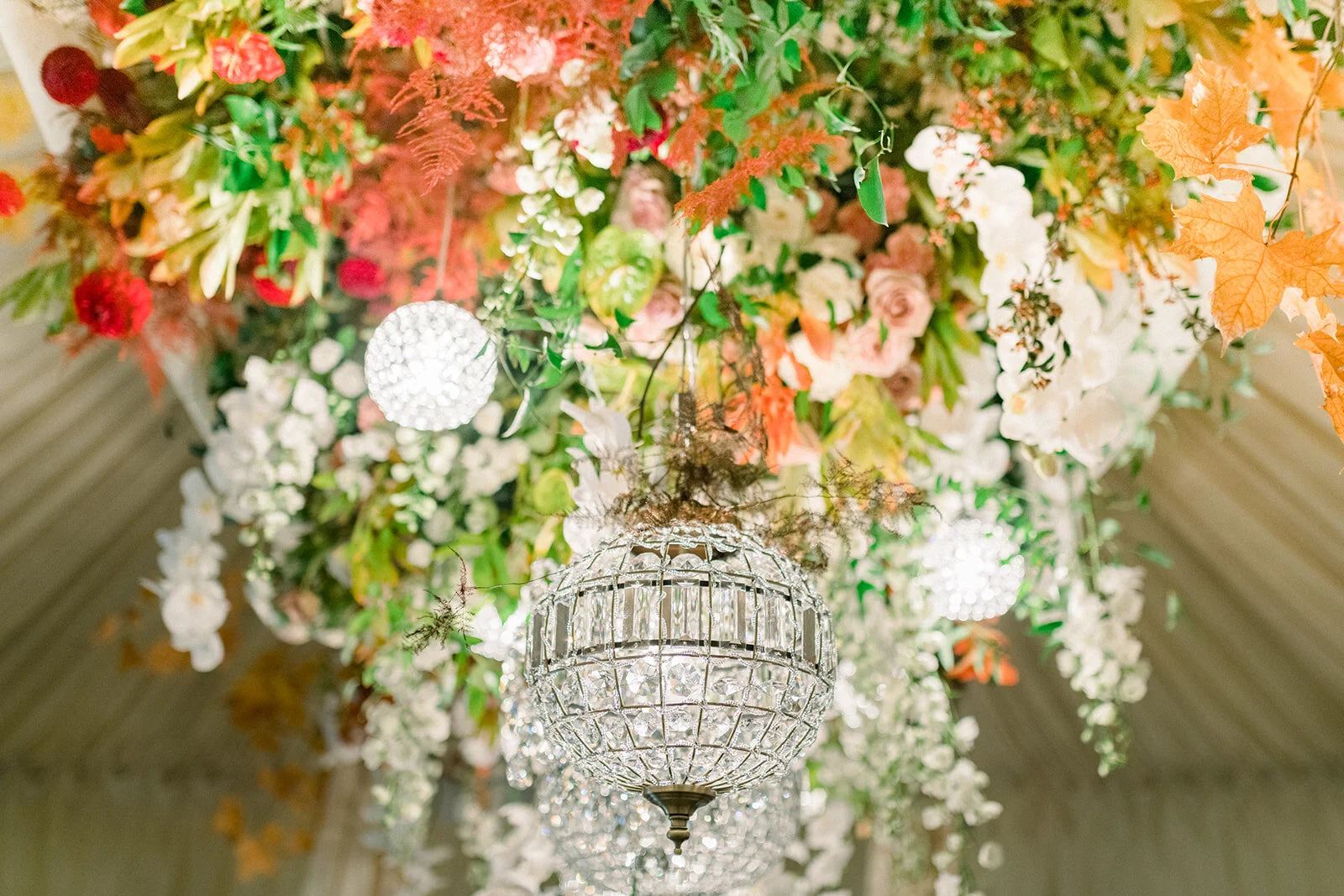 A chandelier hanging from a ceiling decorated with a large arrangement of colorful flowers and autumn leaves.