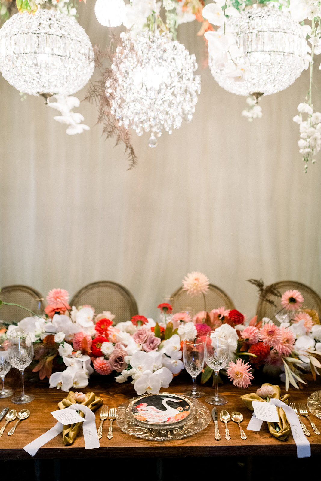 A decorated dining table with a floral centerpiece, wine glasses, gold utensils, and napkins, set against a backdrop of hanging chandeliers and a light-colored curtain.