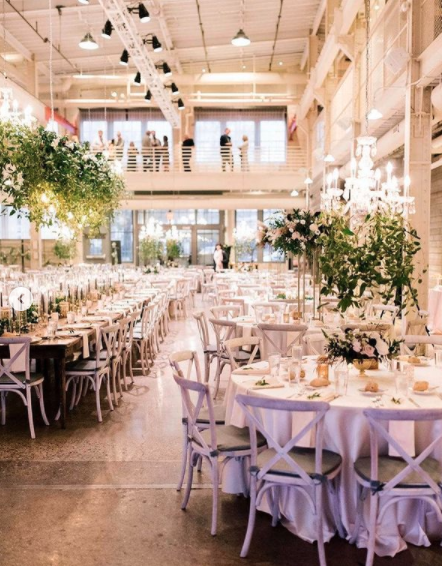 Elegant banquet hall decorated for wedding reception with white tablecloths, floral centerpieces, and chandeliers, featuring a second-floor balcony overlooking the main area.
