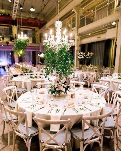 Elegant banquet table decorated with a floral arrangement, candles, and tableware, set in a modern, well-lit event space with chandeliers and multiple round tables.