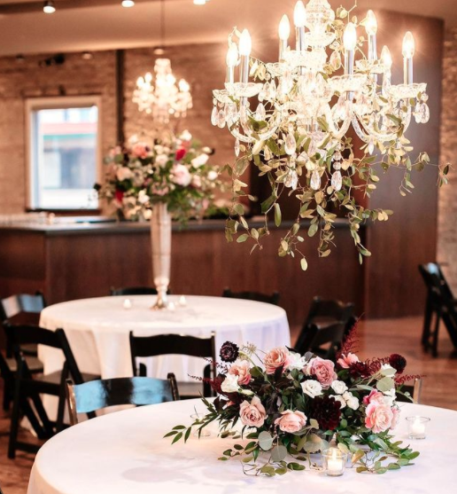 Elegant event space with round tables decorated with floral arrangements and a grand chandelier lighting the room.