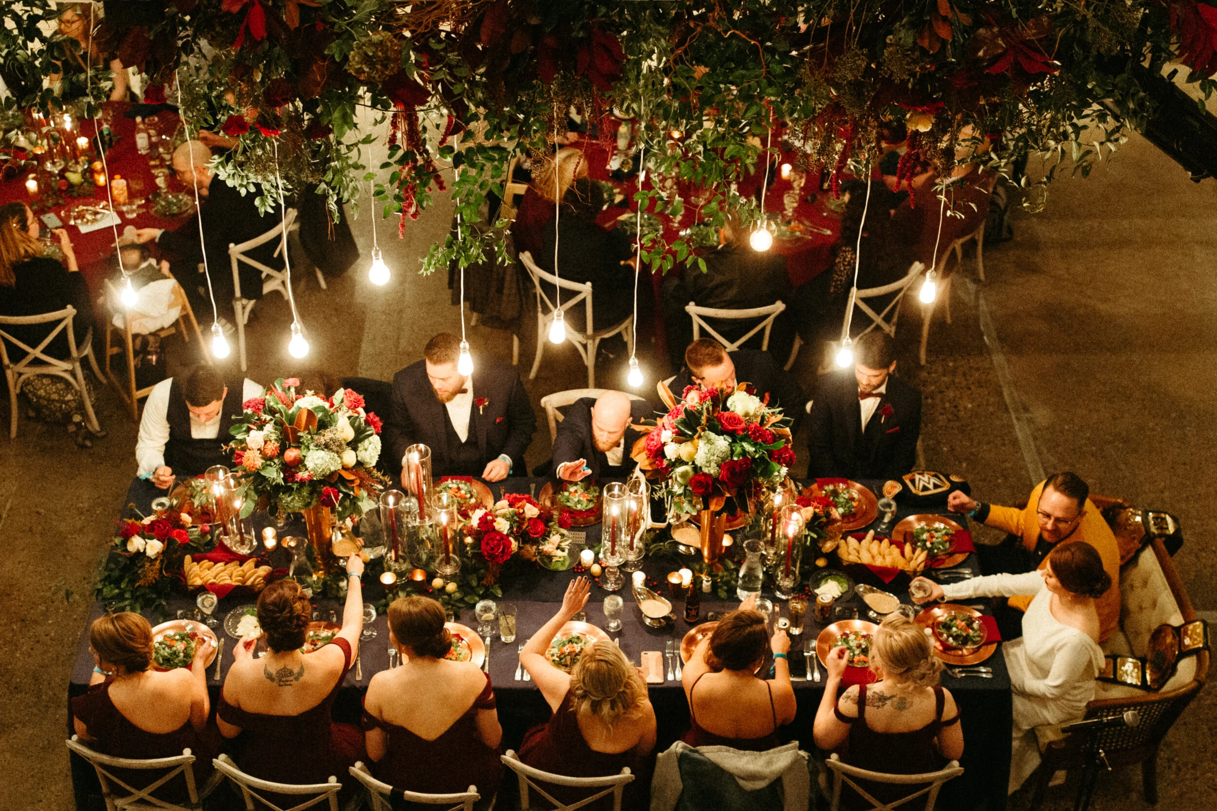 An overhead view of a formal dinner party with a long table decorated with large floral arrangements, candles, and ornate tableware. People are seated around the table, enjoying their meals, in an elegant setting with warm lighting from hanging bulbs