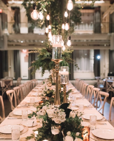 Elegant dining table setup with floral centerpieces and candlelit chandeliers in a rustic indoor venue.