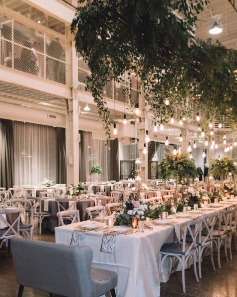 Elegant event space with long tables decorated with white tablecloths, floral centerpieces, and candles. Hanging string lights and greenery add a warm, inviting atmosphere.