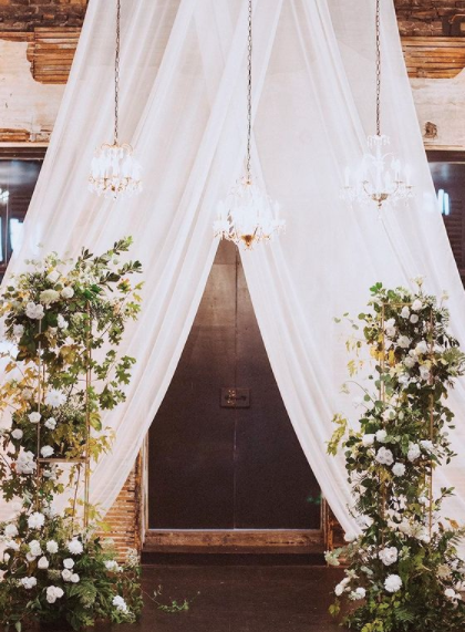Elegant wedding arch with white curtains, hanging chandeliers, and floral arrangements of greenery and white flowers.