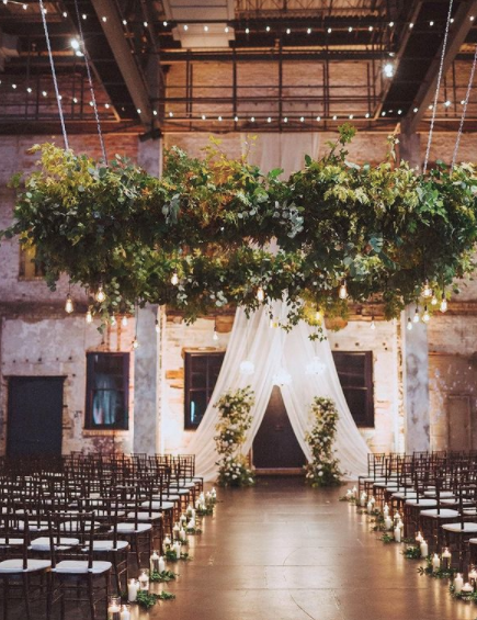Indoor wedding ceremony setup with rows of chairs, floral arrangements, and a draped fabric arch at the altar, with hanging greenery and lights overhead.