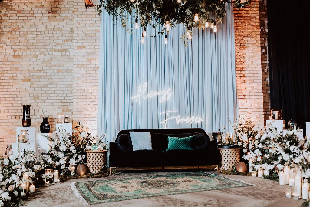 Indoor wedding or event setup with a black couch, decorative pillows, floral arrangements, candles, and a light blue curtain with a neon sign that reads 'Always & Forever.'