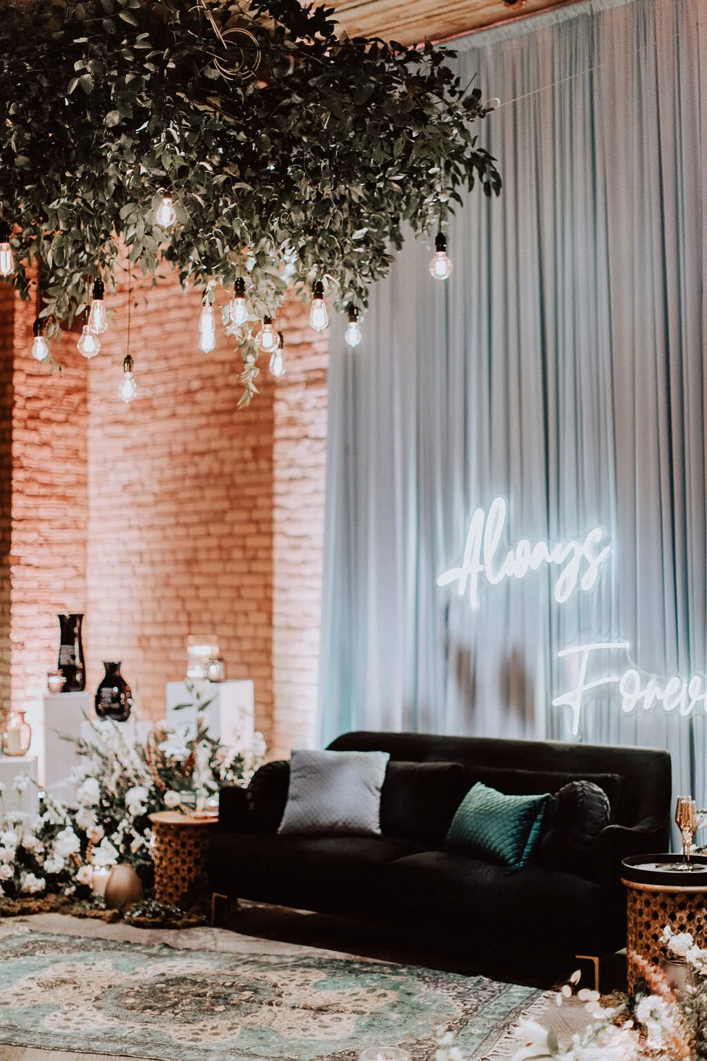 Decorative interior with a black sofa and pillows, a floral arrangement, hanging light bulbs, a neon sign that reads 'Always Forever', and brick wall and curtains in the background.