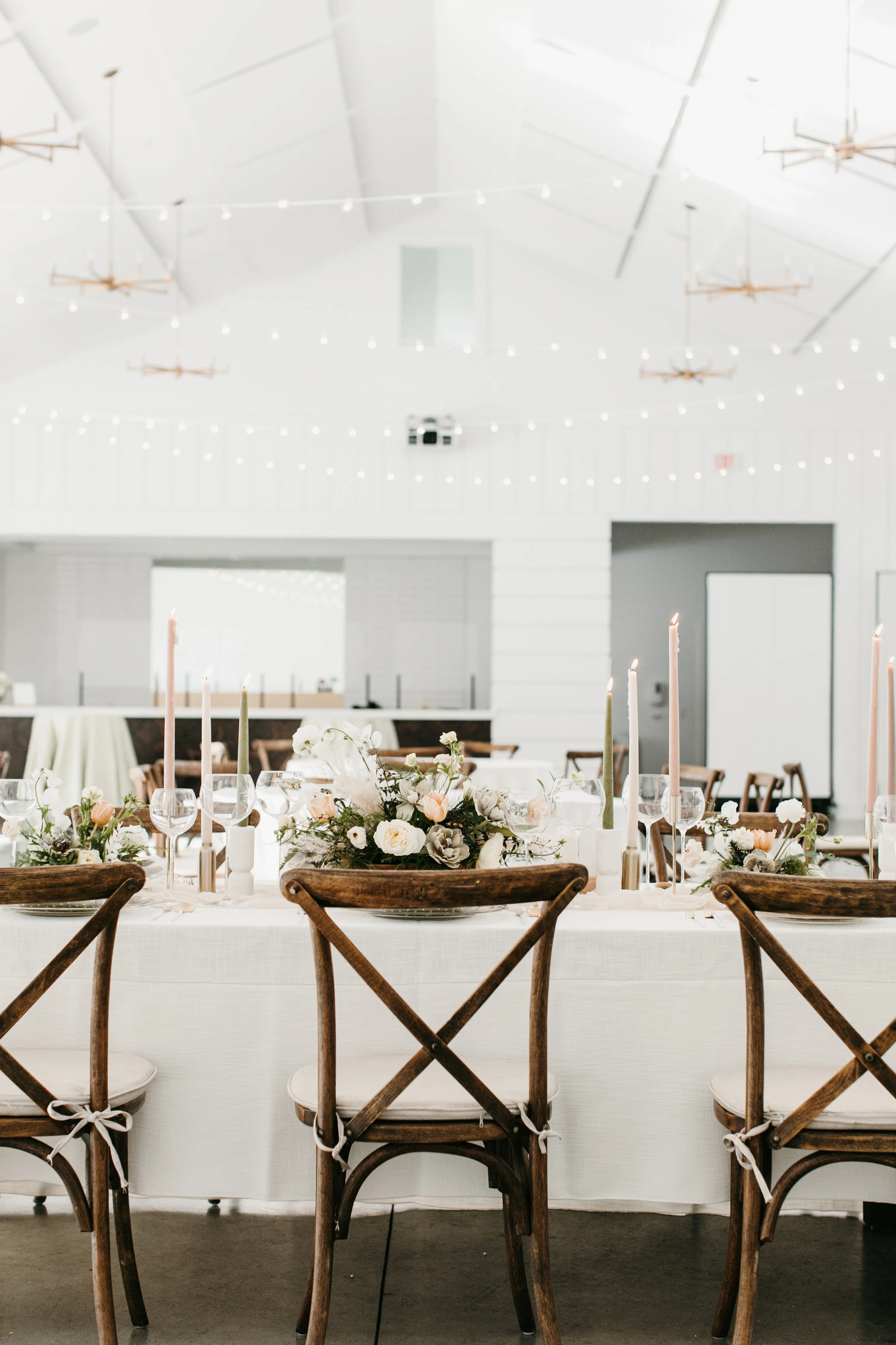 A beautifully decorated wedding reception table with floral centerpieces, candles, and glassware in a bright, modern venue with high vaulted ceilings and string lights.