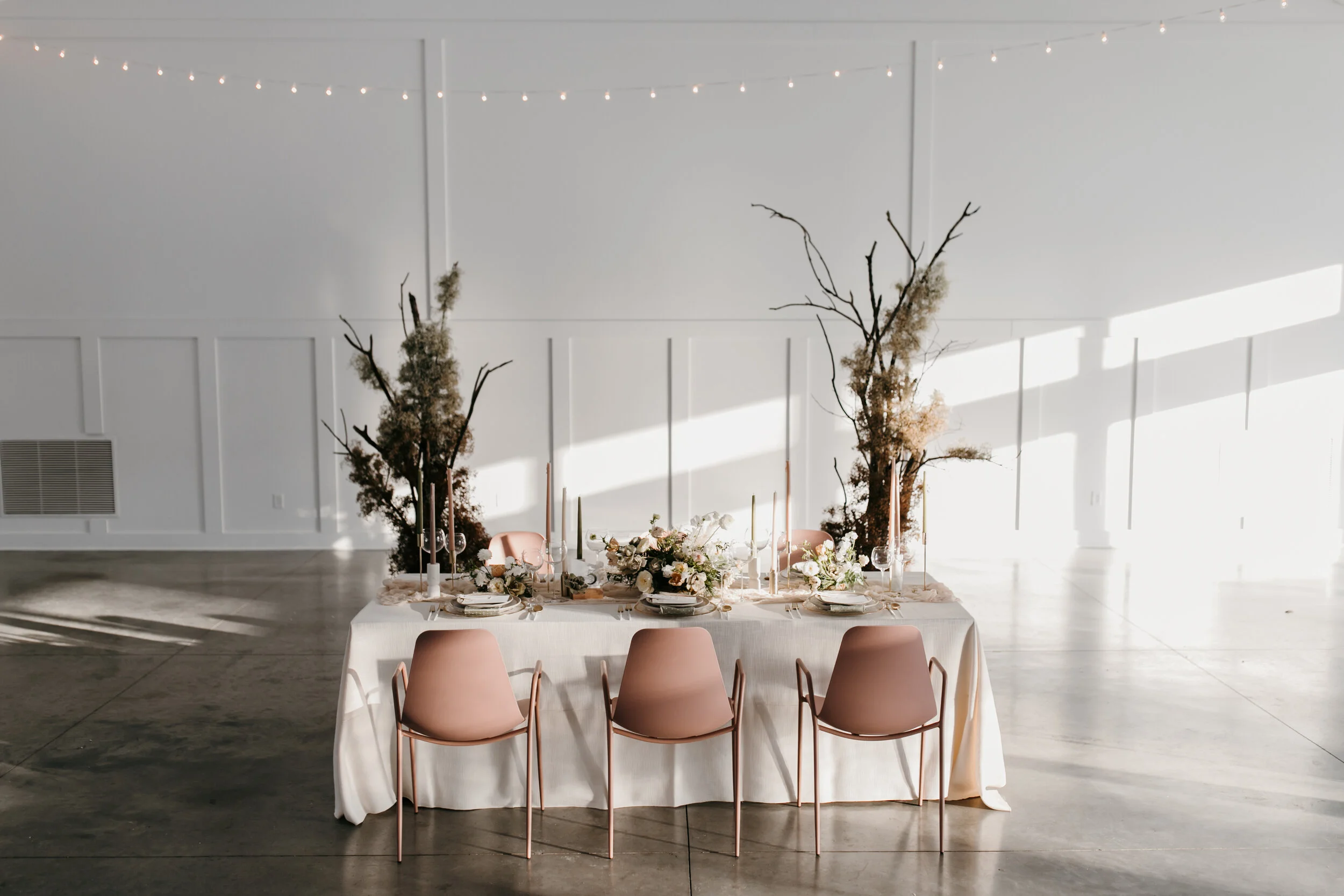 Elegant banquet table decor with pink chairs, floral centerpieces, candles, and natural decorative branches, set in a bright, minimalistic white room with string lights.