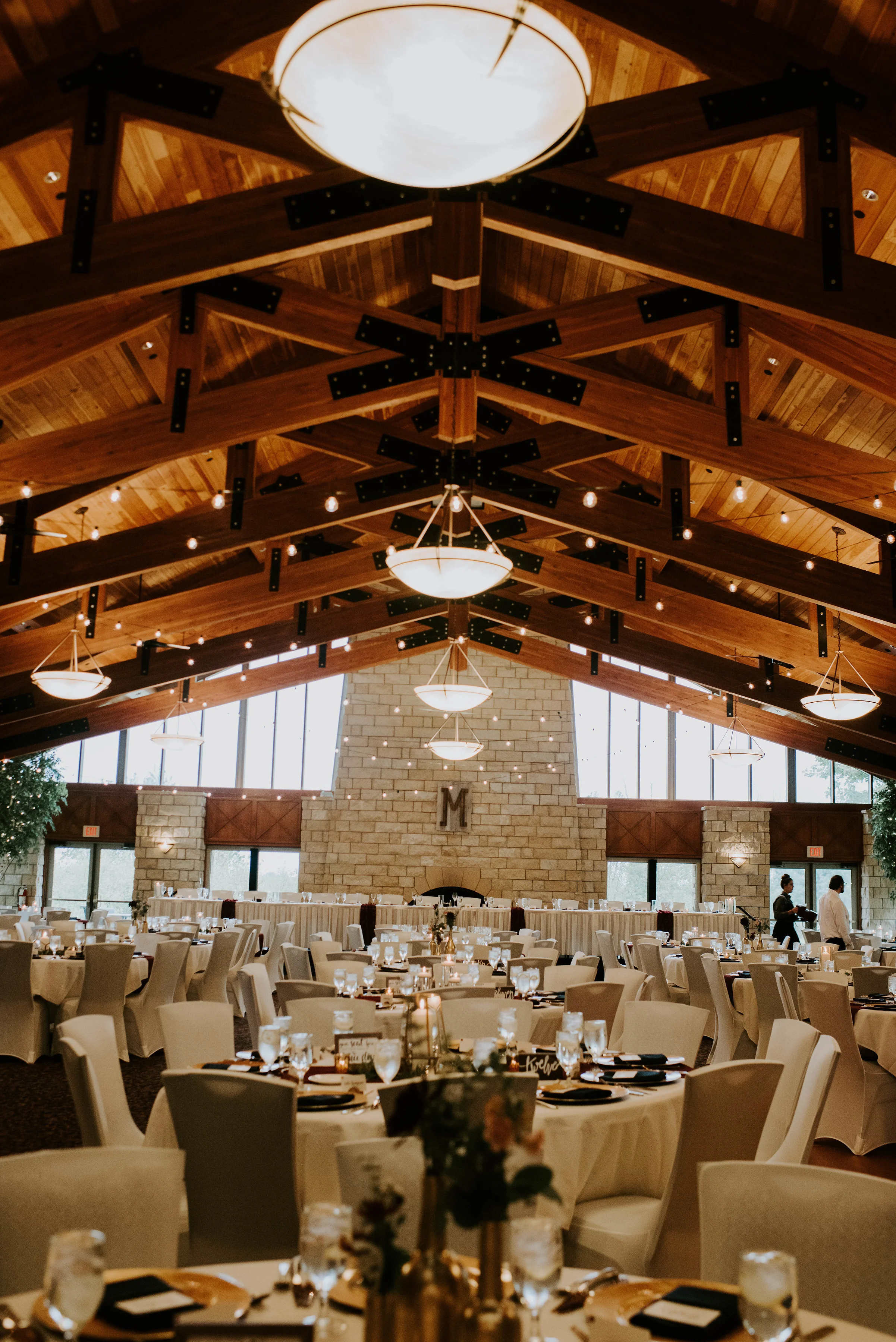 Elegant banquet hall with high vaulted wooden ceiling, hanging light fixtures, and decorated tables with candles and floral centerpieces, set for a formal event.