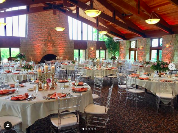 Elegant banquet hall with round tables set for a formal event, decorated with pink and white floral centerpieces, candles, and salmon-colored napkins. The room features large windows, a brick fireplace, and exposed wooden beams on the ceiling.