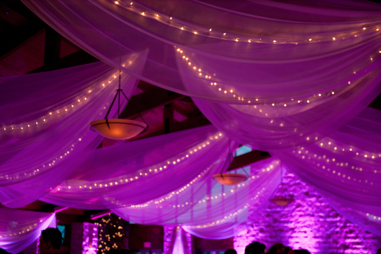 Indoor event space decorated with purple drapery and string lights, with hanging light fixtures and a brick wall illuminated with purple lighting.