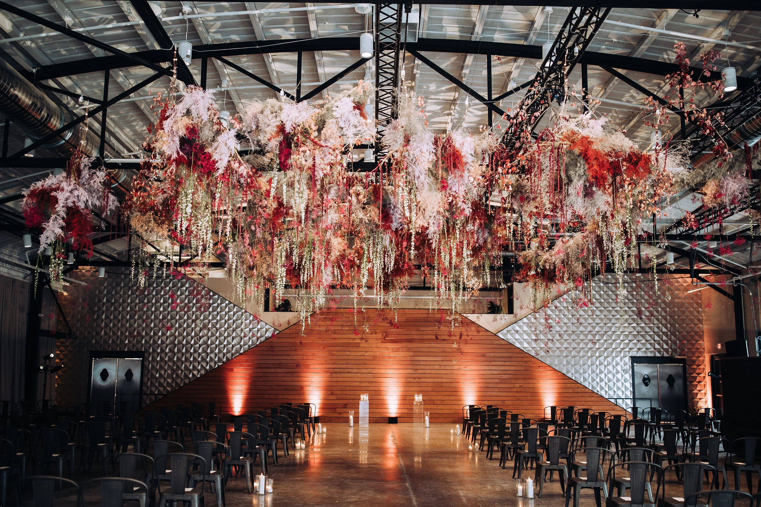 Interior of an event space decorated with a large, elaborate floral installation hanging from the ceiling, featuring pink, white, and red flowers and greenery. The room has a wooden stage, surrounded by black chairs, with candles placed along the ais