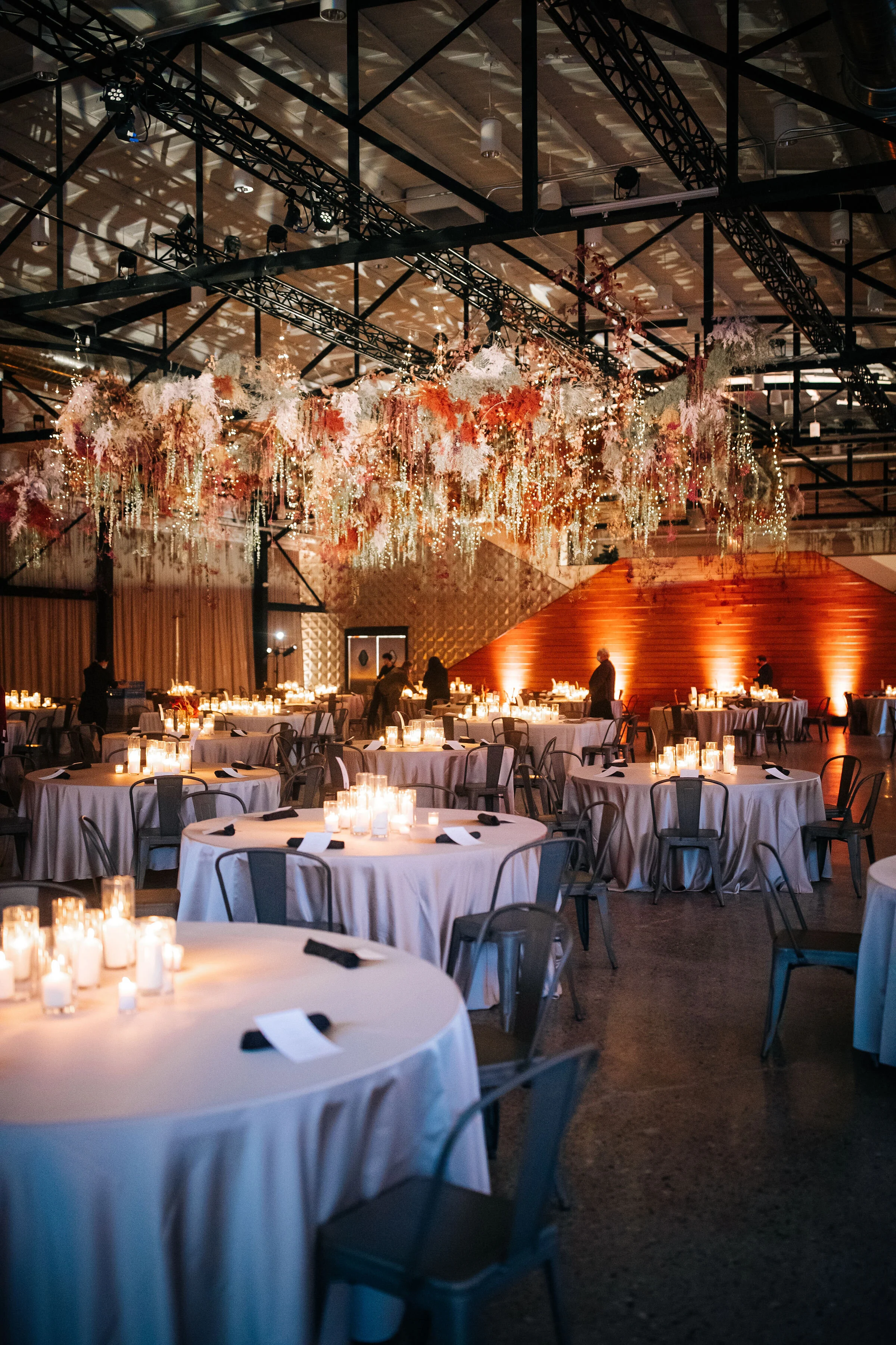 Elegant event space decorated with floral arrangements hanging from the ceiling and lit with candles on round tables covered with white tablecloths, set for a formal occasion.