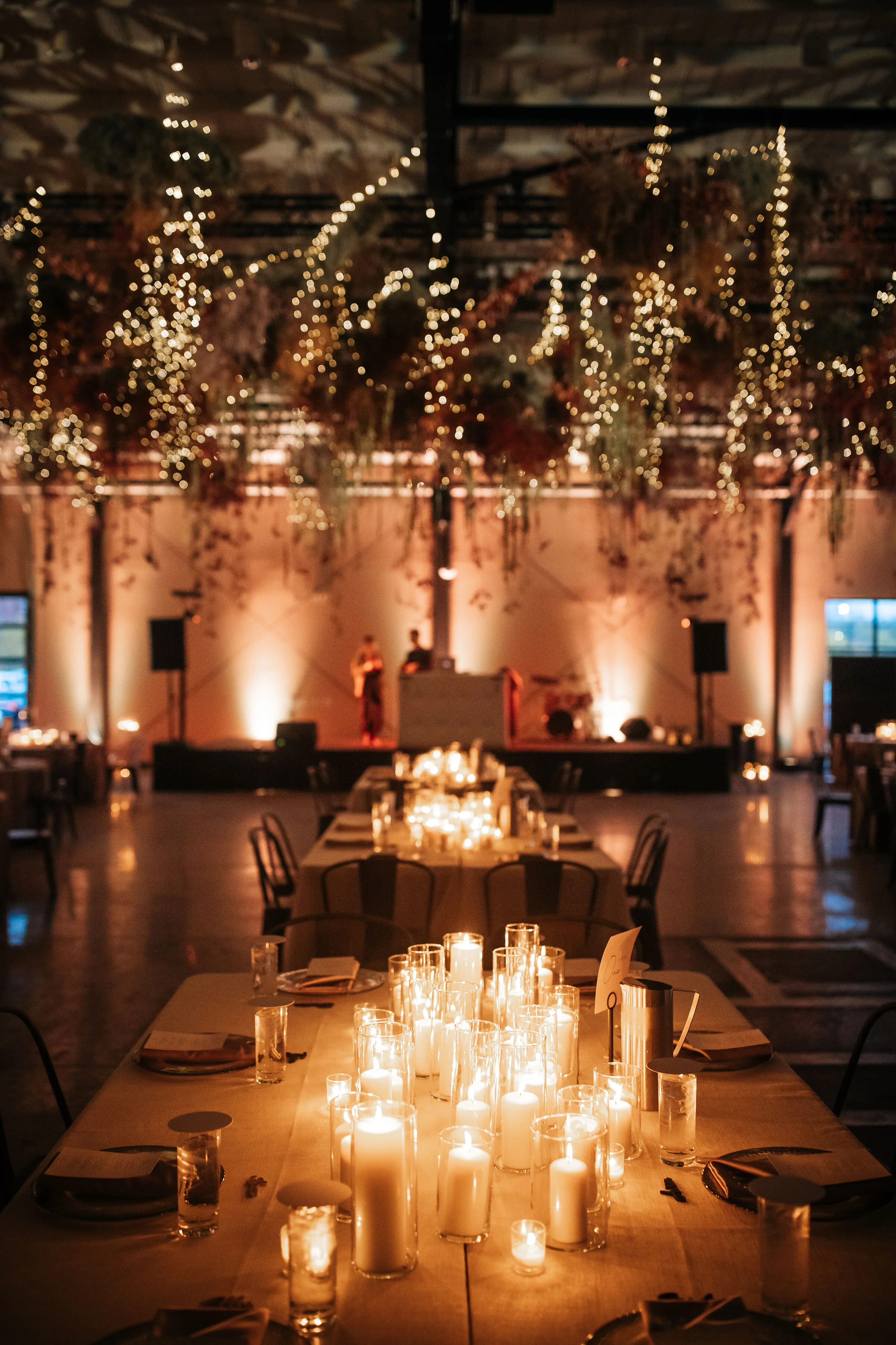 A dimly lit event space or banquet hall decorated with candles on tables, hanging string lights from the ceiling, and soft uplighting on the walls, with a stage in the background where a performer is singing.