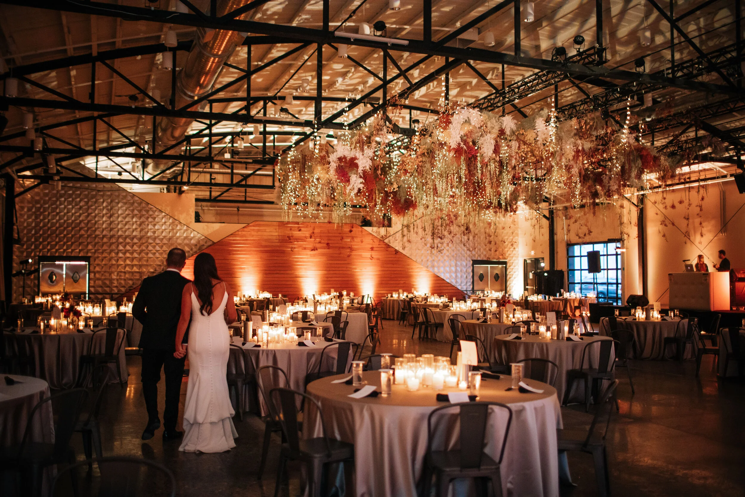 Indoor wedding reception with round tables, candle centerpieces, and hanging floral decorations in warm lighting.