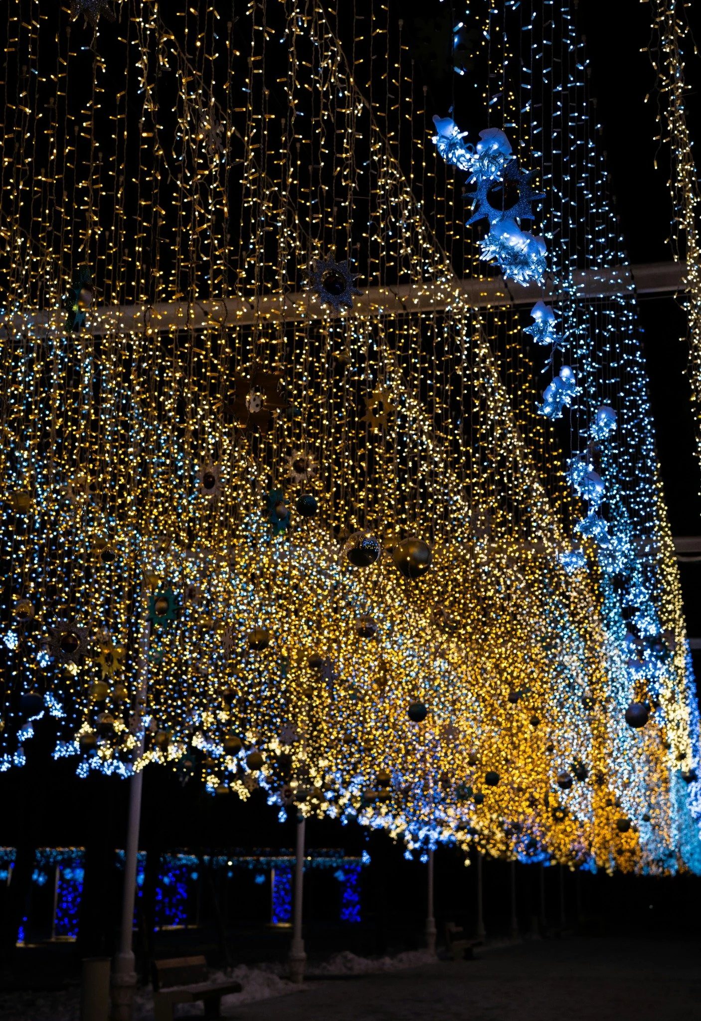 1. Dense canopy of warm gold and cool blue holiday lights suspended overhead with ornaments and snowflake accents.