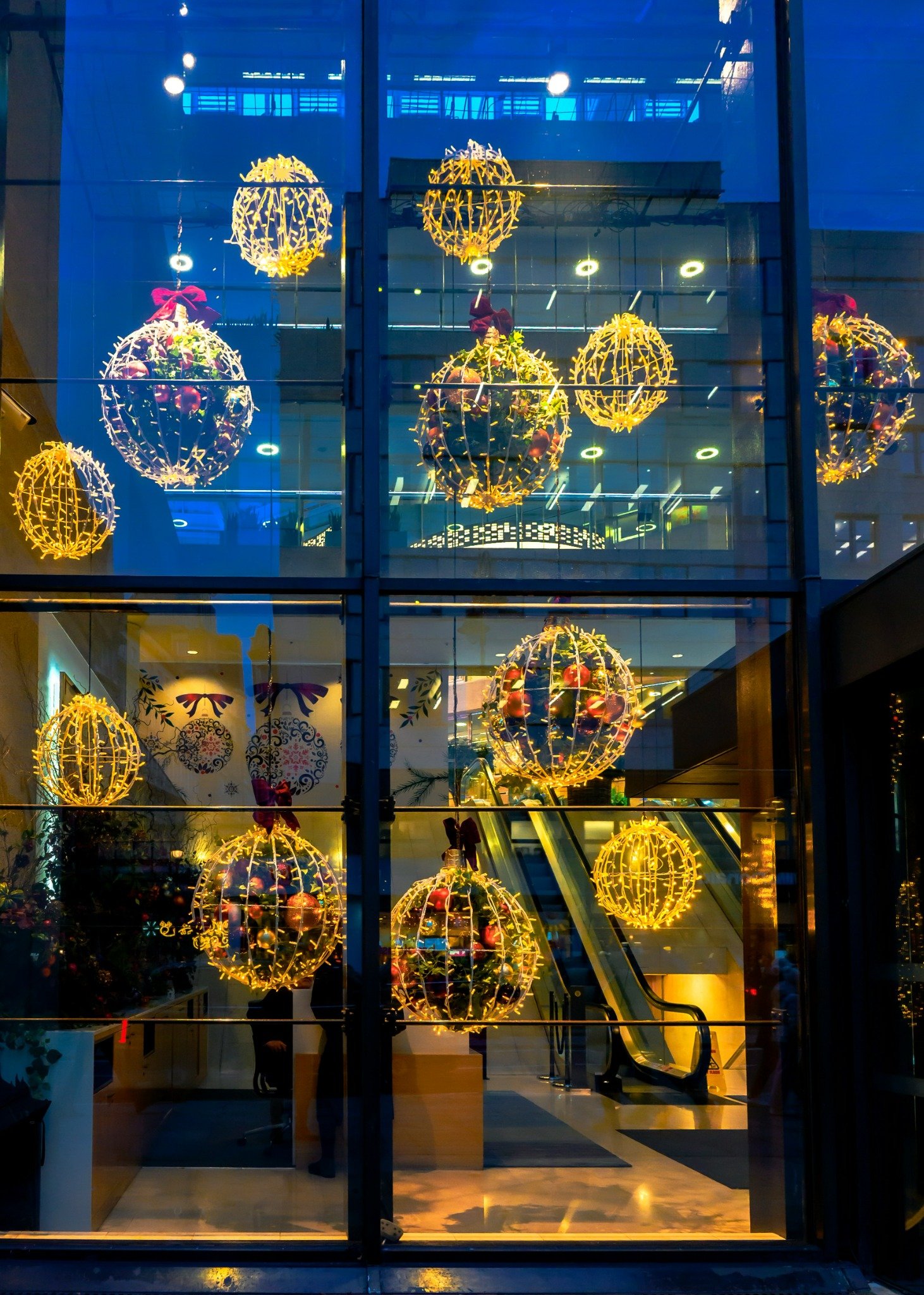 Oversized illuminated holiday ornaments suspended inside a multi-story glass commercial entrance.