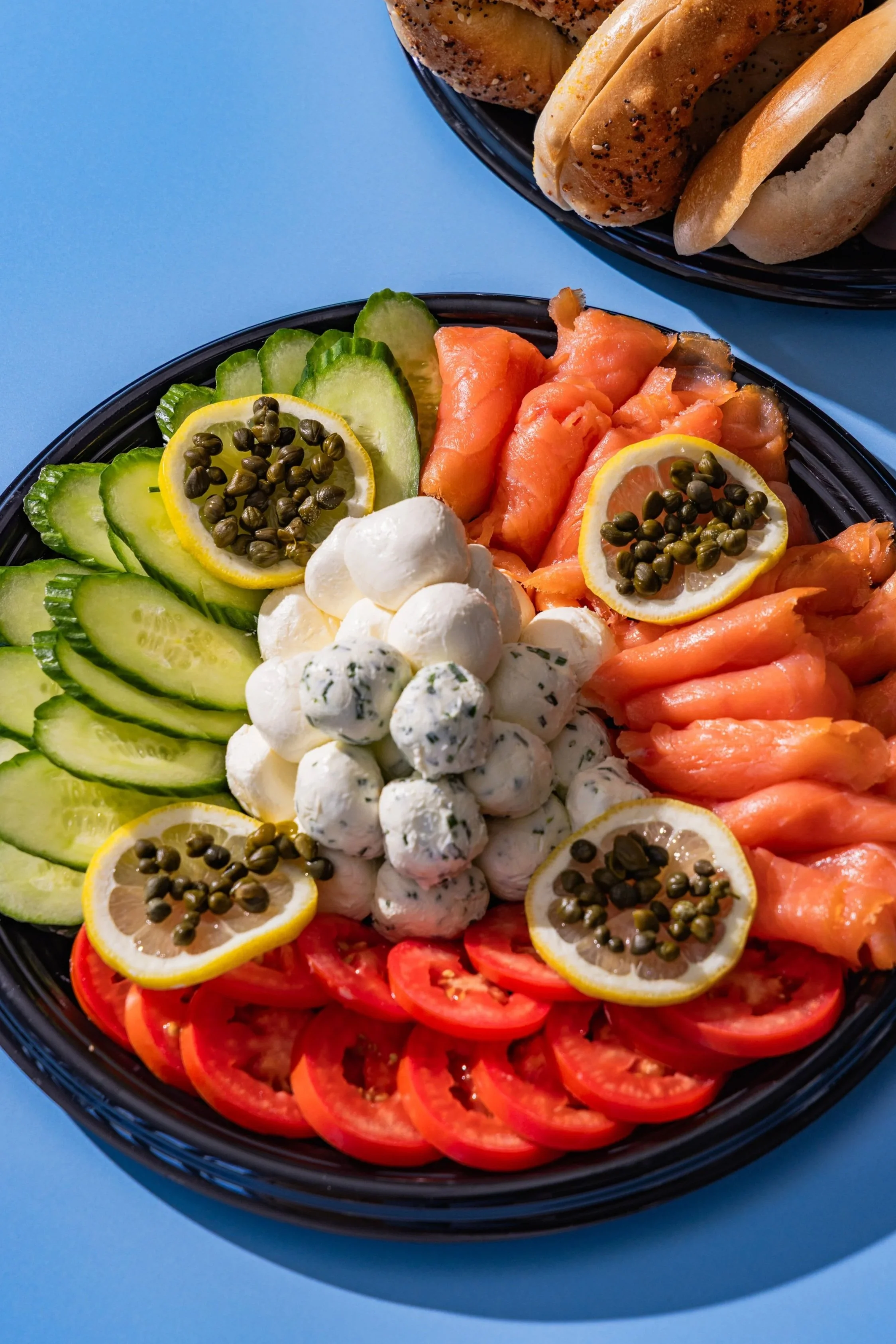 Colorful platter of sliced cucumbers, salmon, cherry tomatoes, lemon slices topped with capers, and small mozzarella balls, with a basket of bread rolls in the background on a blue surface.