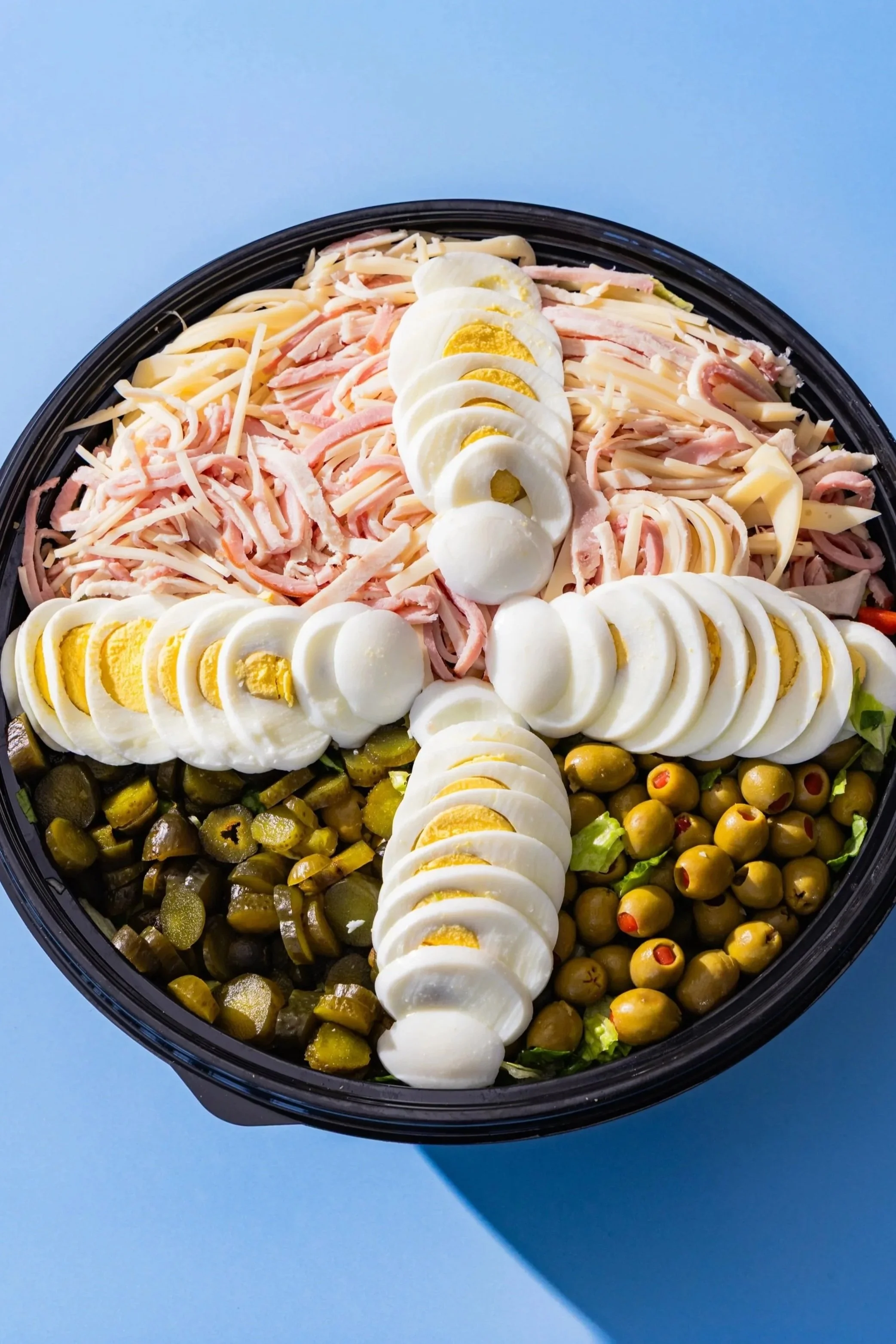 A black container filled with a salad consisting of sliced hard-boiled eggs, shredded ham, shredded cheese, green olives with red pimentos, and chopped lettuce, set against a blue background.