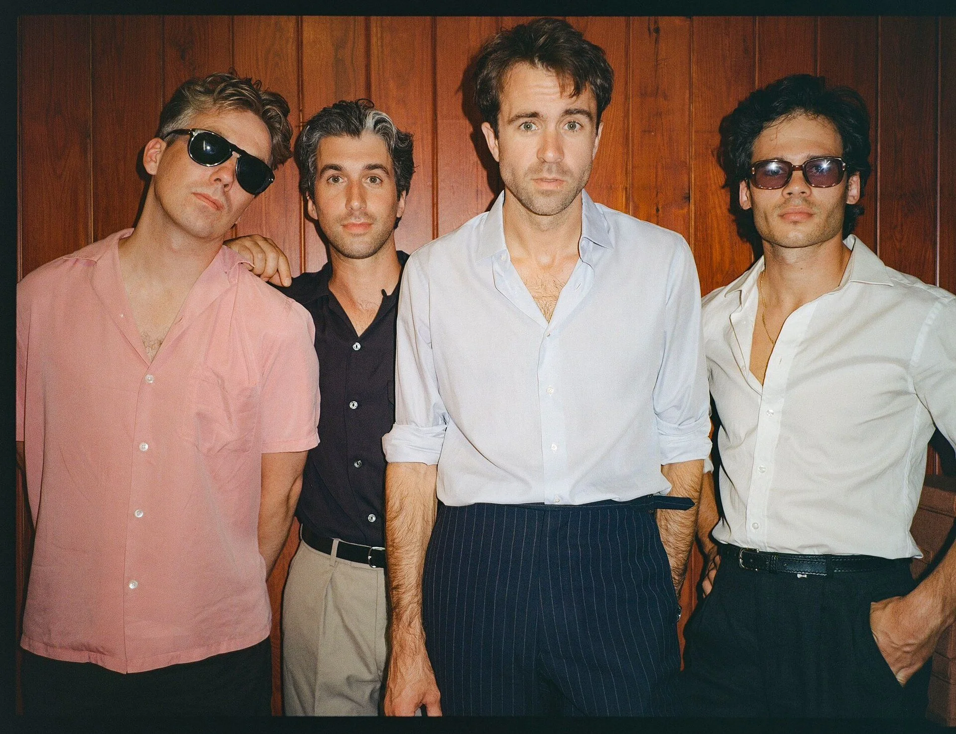 Four men standing in front of a wooden wall, wearing casual button-up shirts and sunglasses.