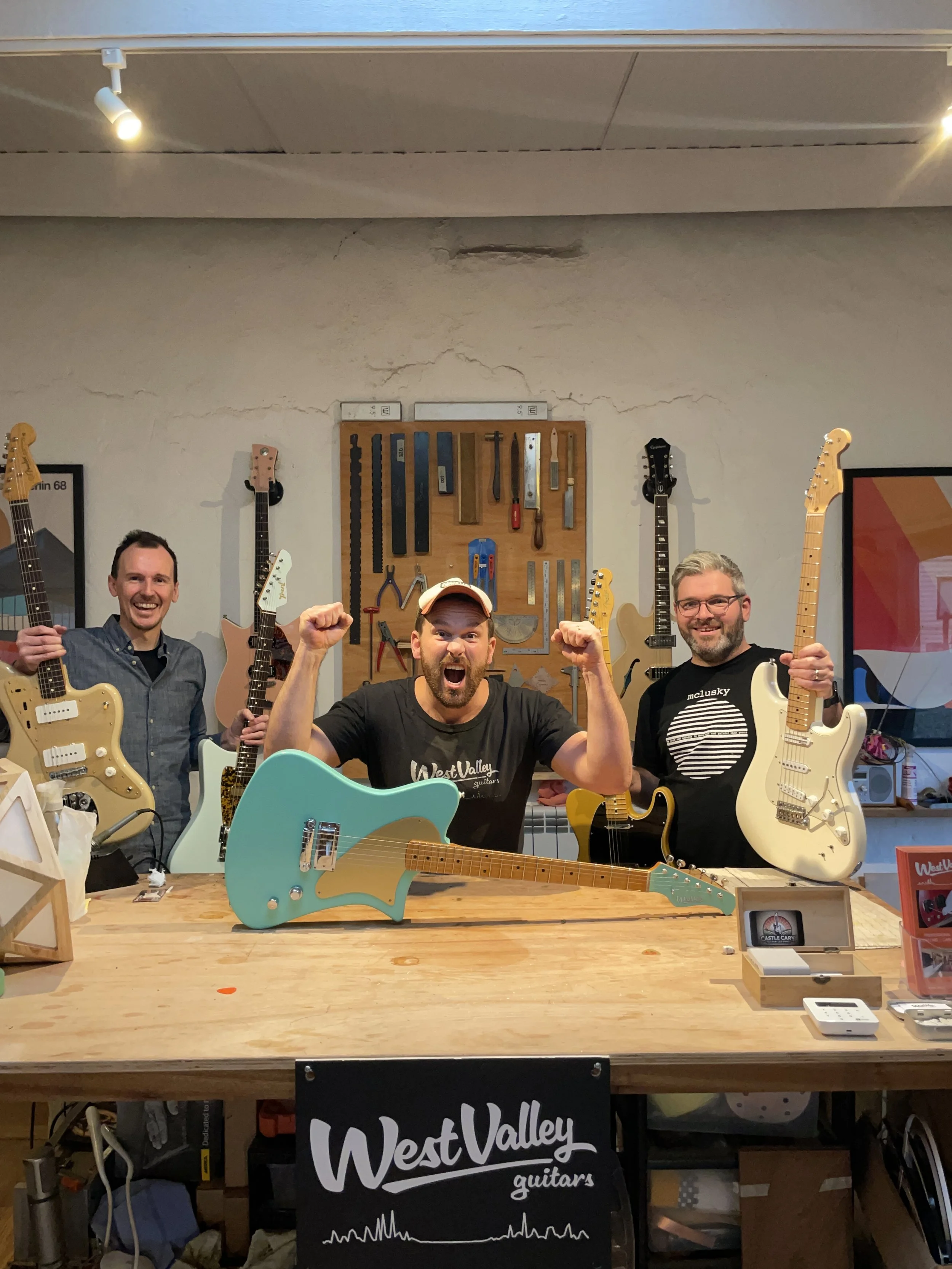 Three men standing behind a workbench with electric guitars, smiling. The middle man is raising his fists in excitement. The workspace has guitar tools mounted on the wall in the background, and a "West Valley Guitars" sign is displayed on the bench.