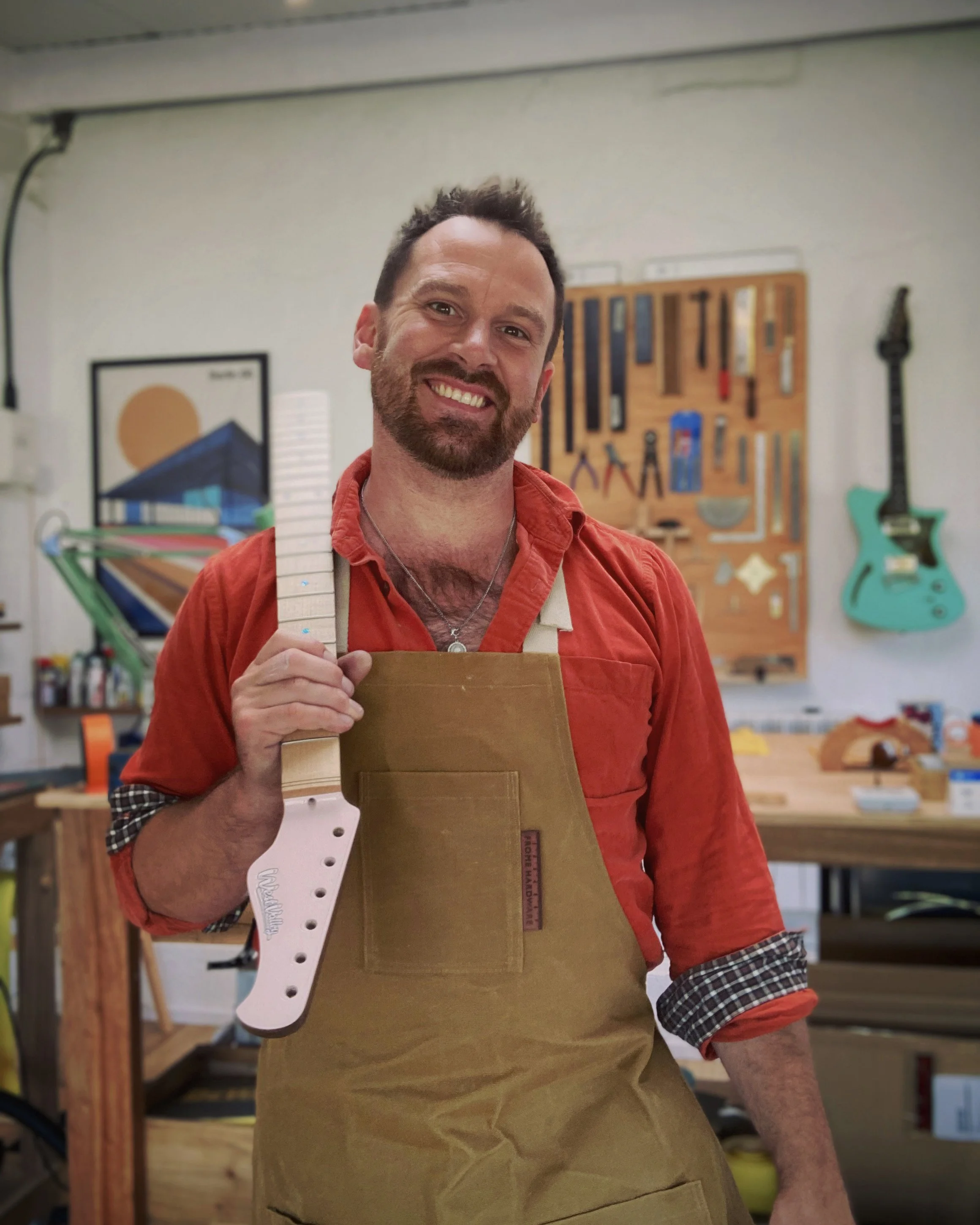 Person in apron holding a guitar neck in workshop with tools on wall and guitar behind.
