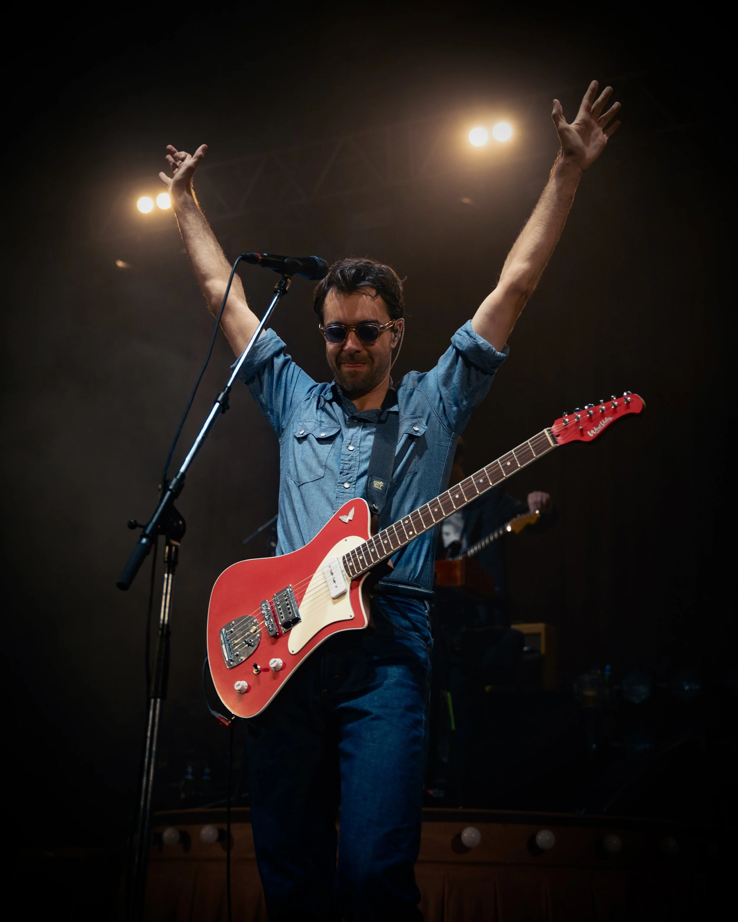 Musician with red electric guitar on stage, arms raised, wearing sunglasses and denim shirt, under stage lights.