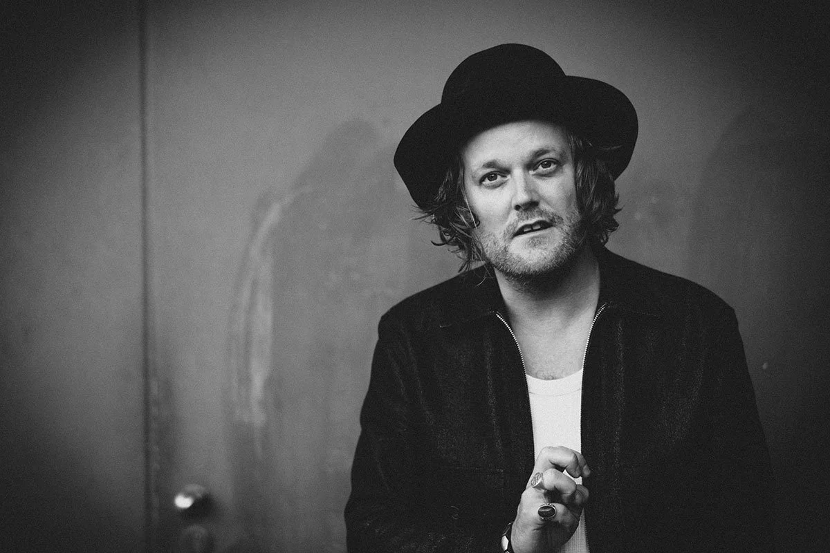 Black and white portrait of a man wearing a hat and jacket, looking at the camera, possibly holding an object in his hand.