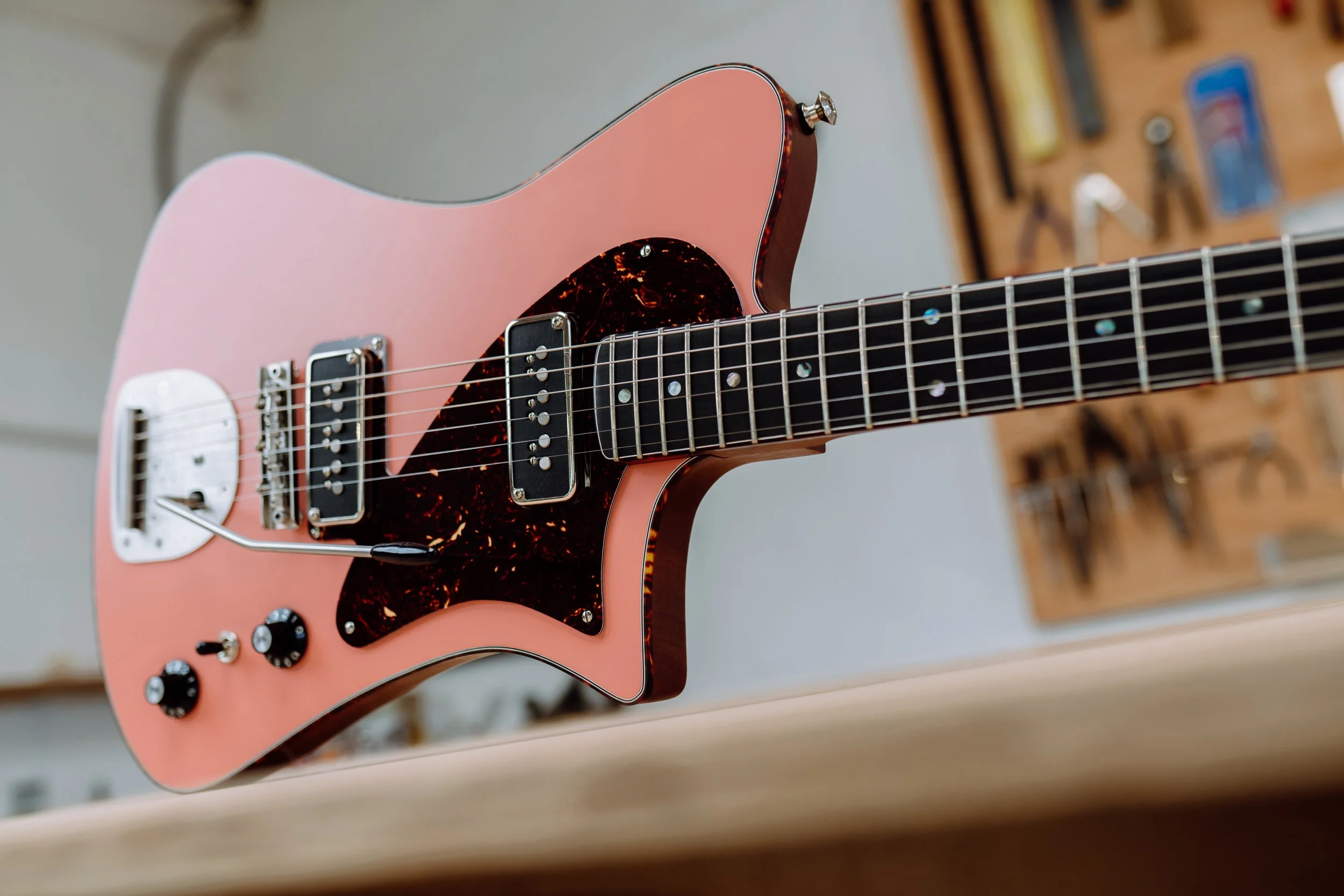 Close-up of a pink electric guitar with black pickguard and knobs, resting on a wooden surface in a workshop setting.