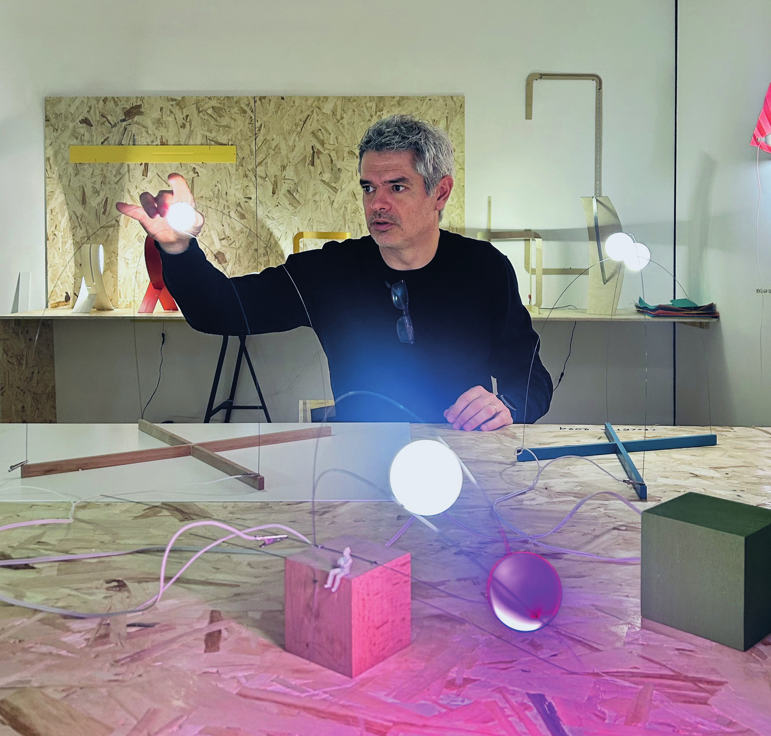 A man with gray hair and a black shirt interacts with illuminated orbs and wires on a table in a modern, minimalistic workspace with geometric and wooden elements.