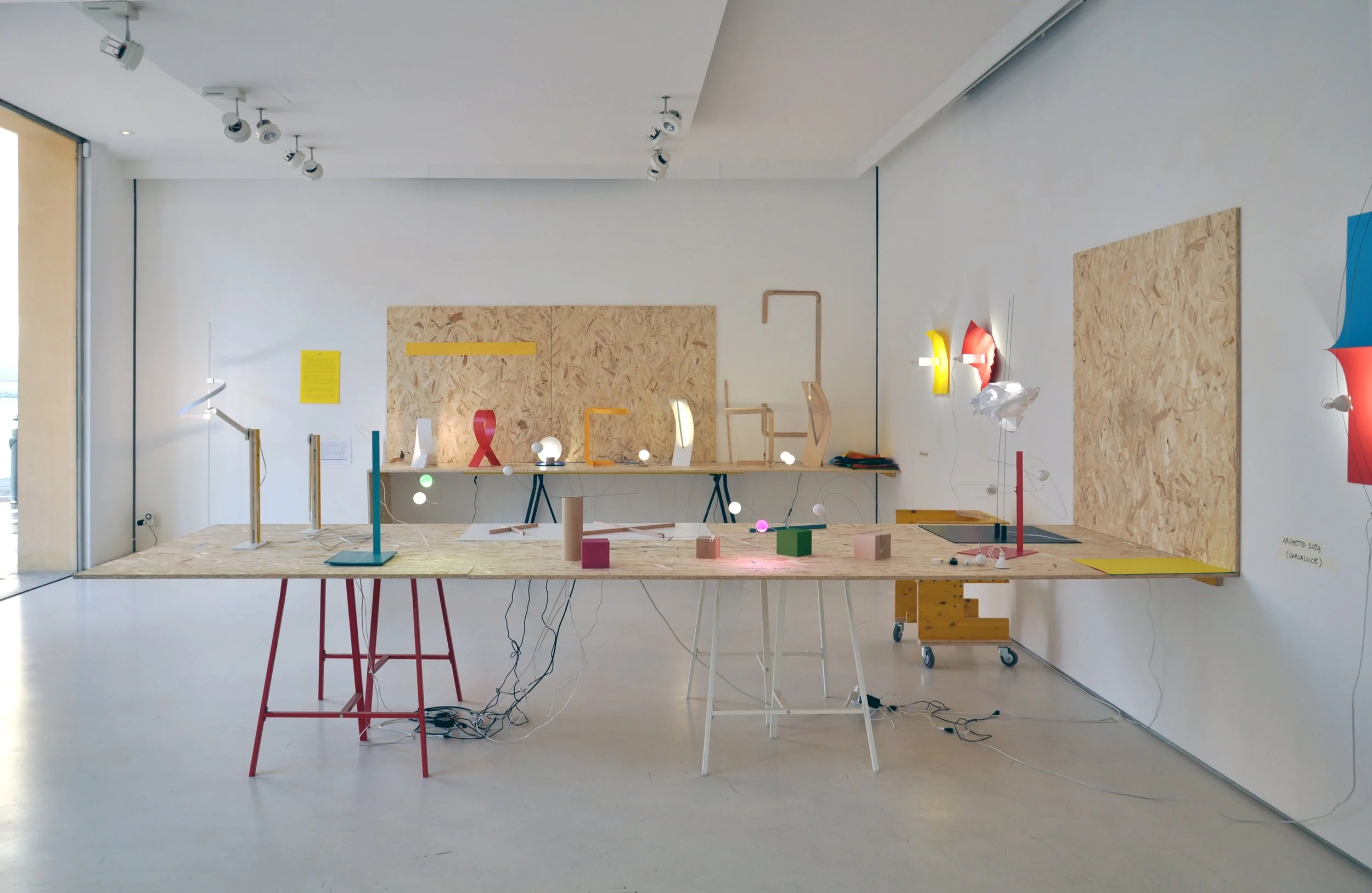 Art installation or design studio with various colorful, abstract sculptures and models on plywood tables and boards in a white-walled room.