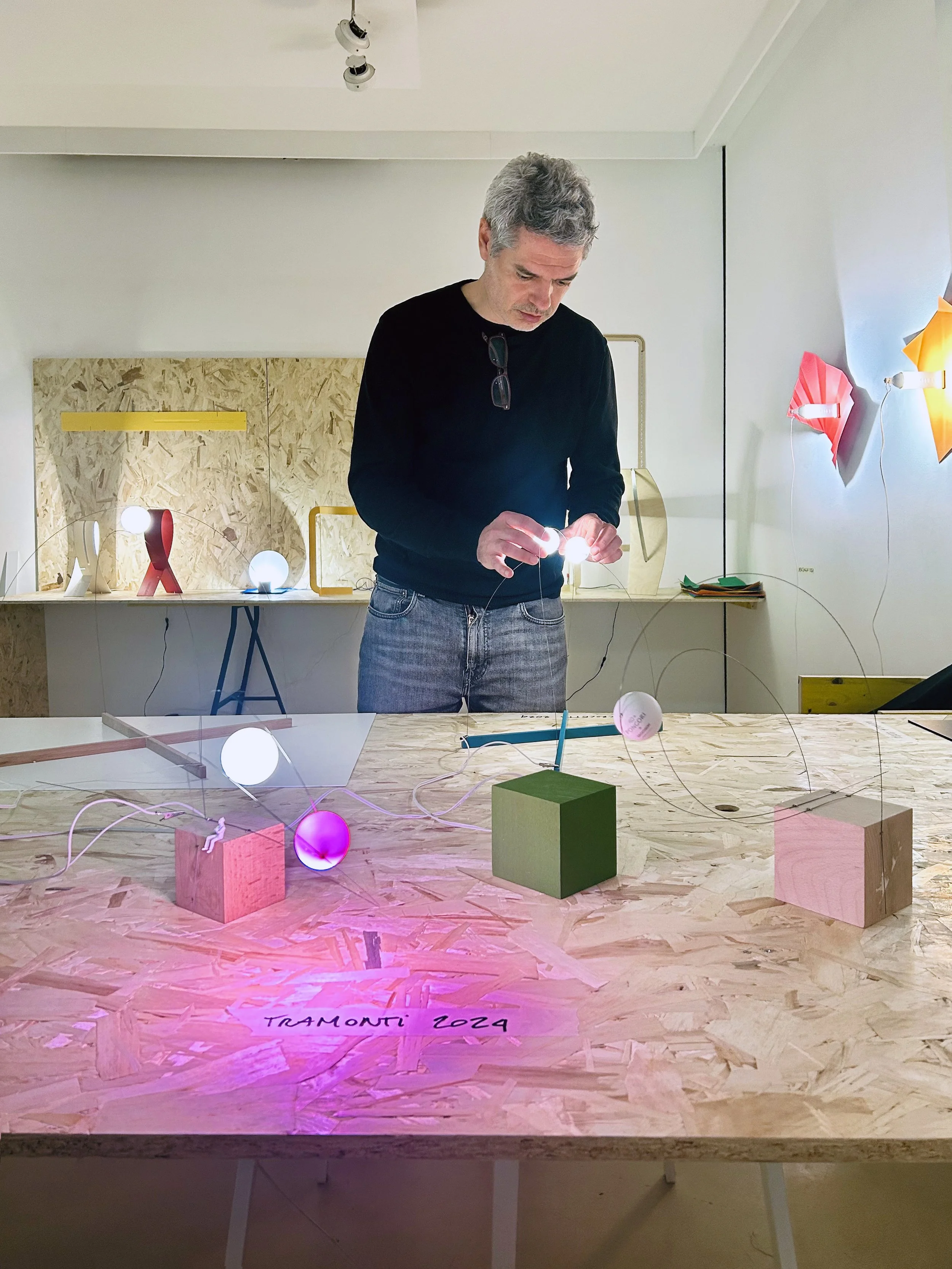 A man adjusting a small illuminated object on a table in an art or design studio, with similar glowing objects and geometric shapes on the table and in the background.