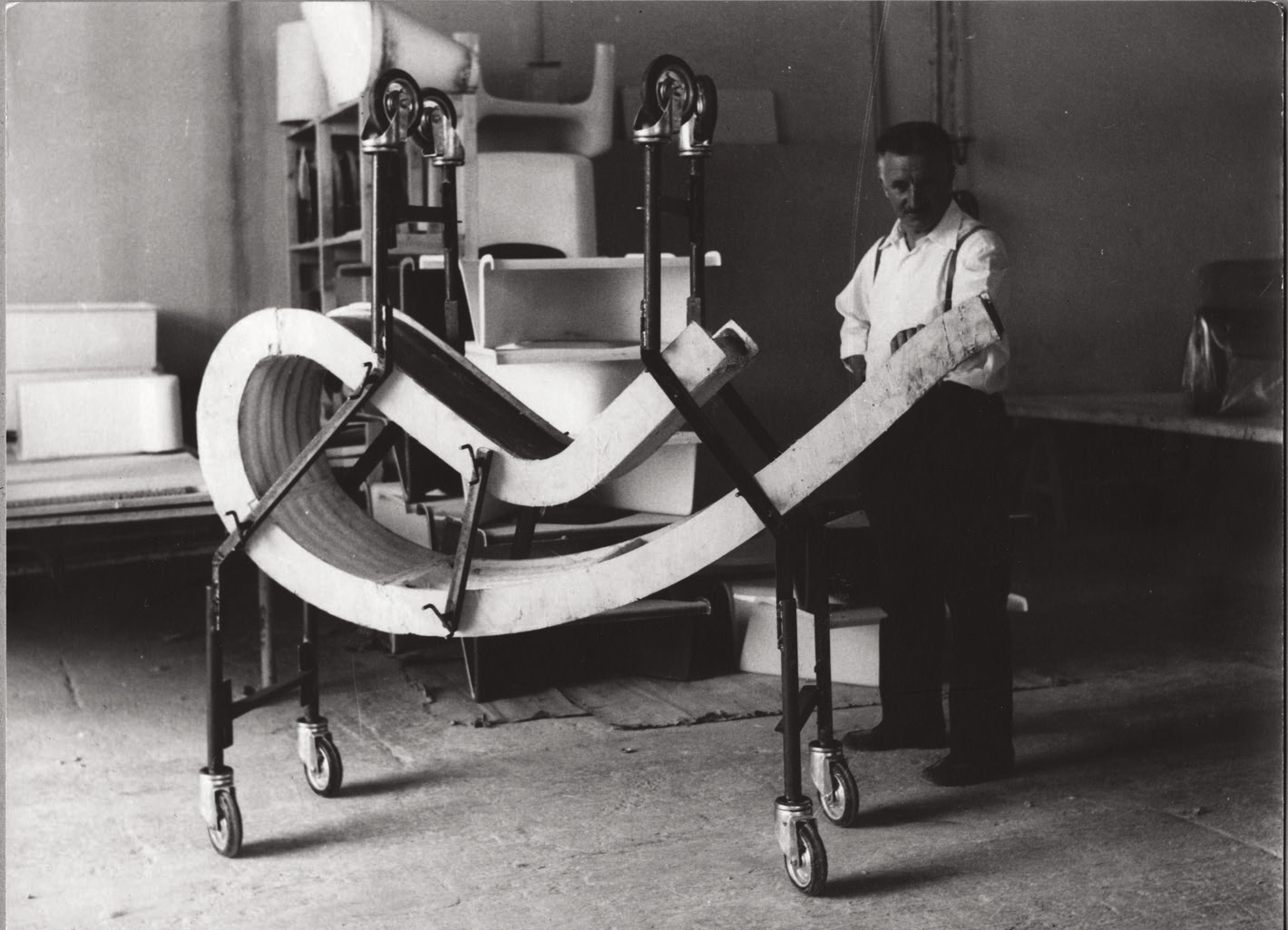 A man in a white shirt stands beside a large mechanical sculpture in a workshop or factory setting. The sculpture has a curving, tube-like structure supported by a wheeled frame.