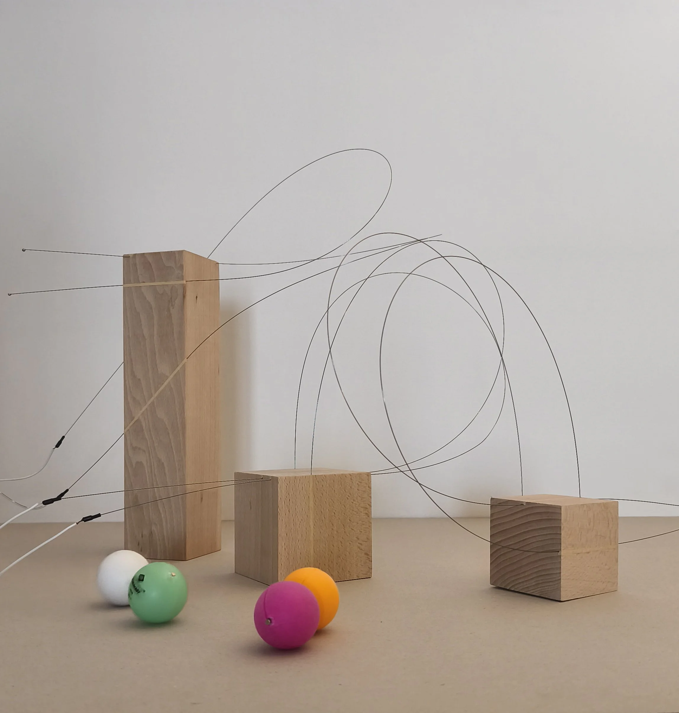 Abstract art installation featuring three wooden blocks of varying heights, thin wire loops extending from two of the blocks, and four colorful foam balls on a beige surface against a plain white wall.