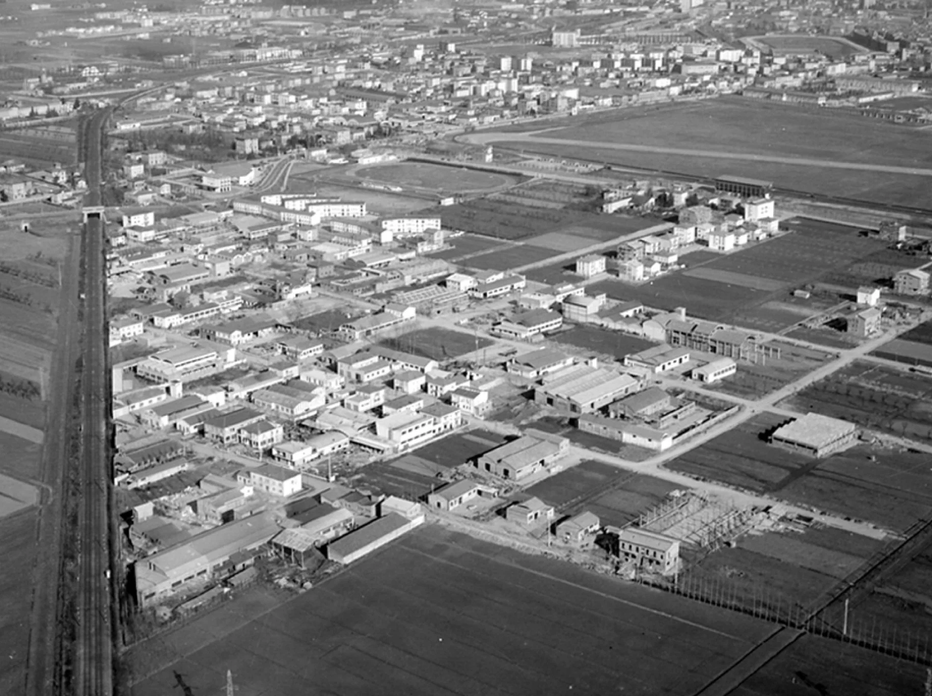 Aerial black and white photo of a city neighborhood with houses, streets, and train tracks, with farmland surrounding the area.