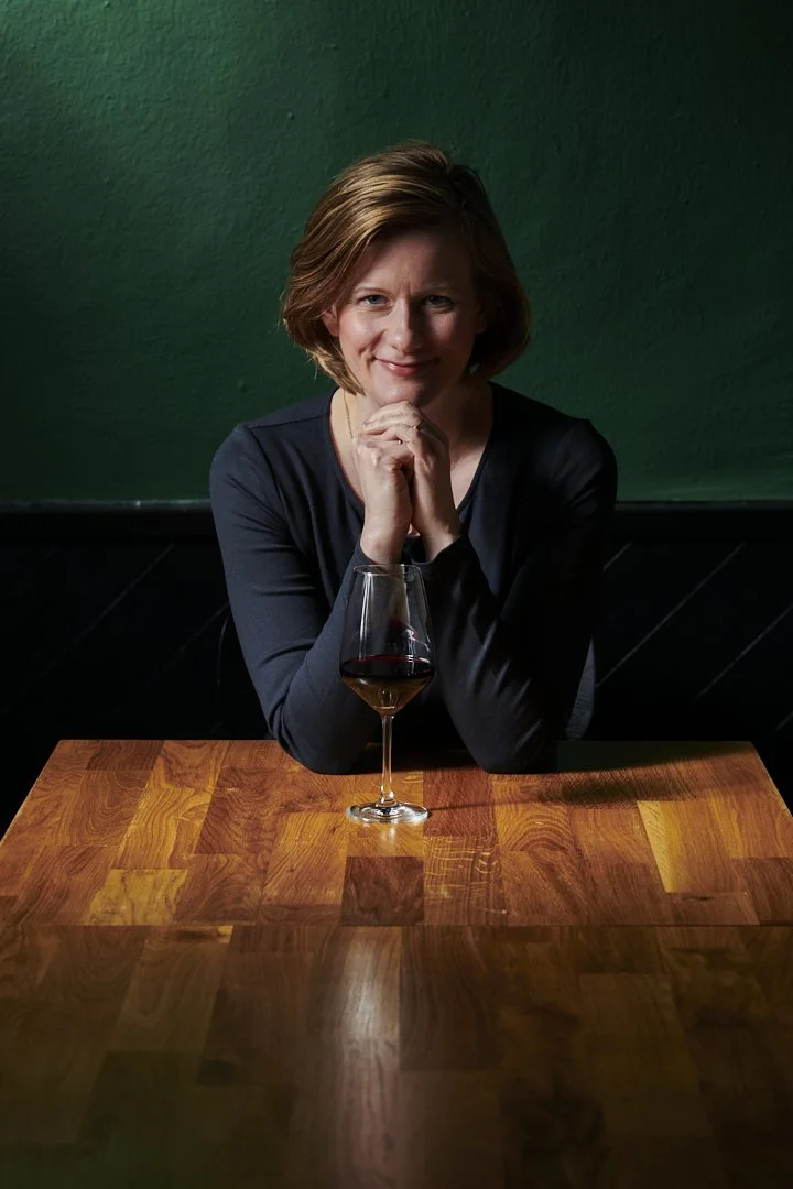 Kristina sitzt an einem Holztisch mit einem Glas Rotwein darauf, gestrichener grüner Hintergrund.