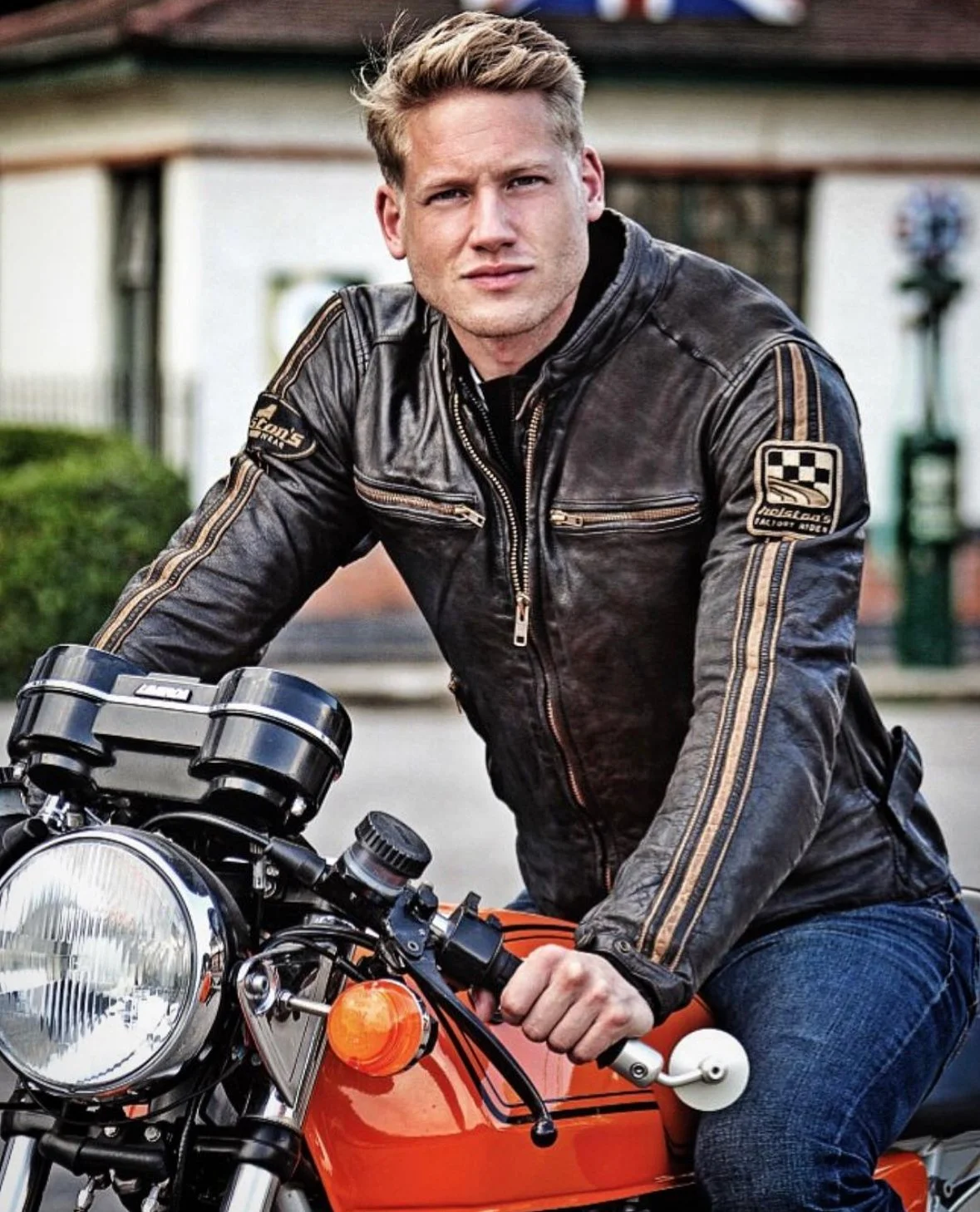 A young man with blonde hair wearing a black leather jacket and blue jeans, sitting on an orange motorcycle outdoors.