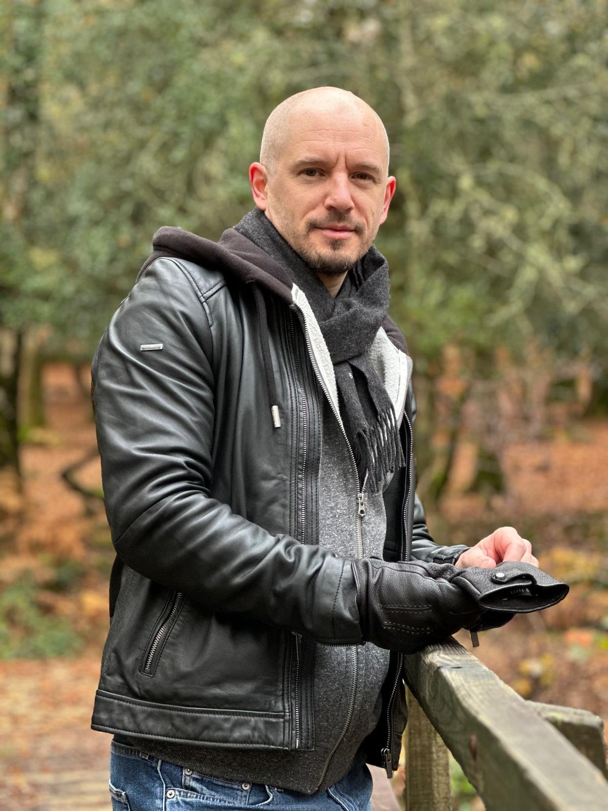 A man standing outdoors on a wooden bridge in a forest during fall, wearing a leather jacket, scarf, gloves, and hoodie.