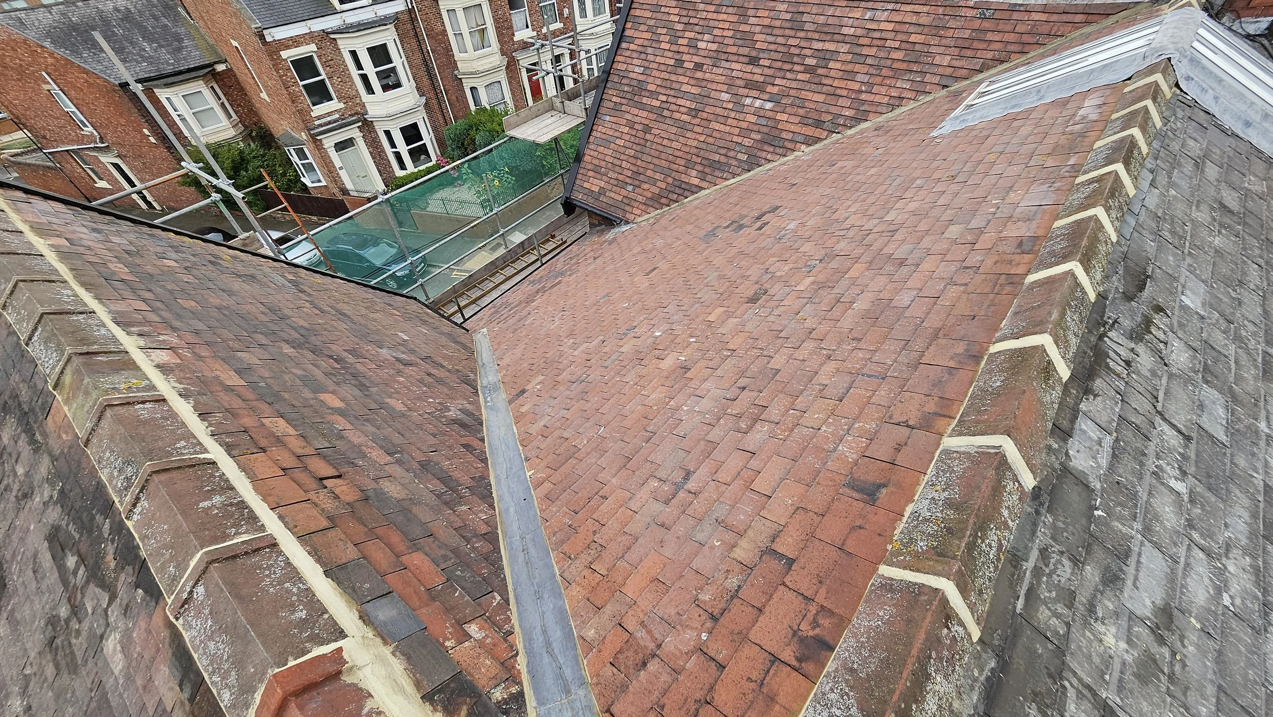 View from a rooftop showing multiple rooftops covered with red bricks, part of a scaffolding structure, and a street with residential houses and a parked car.