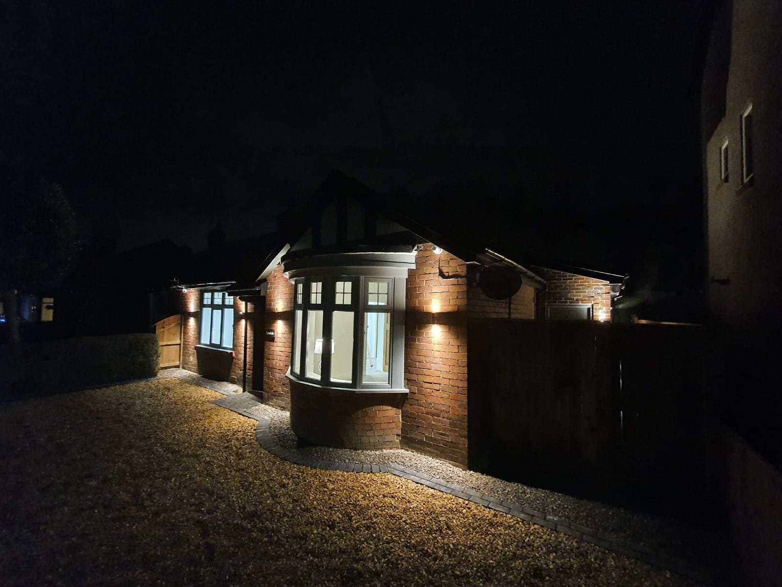 Night view of a brick house with illuminated porch and bay window, surrounded by a gravel yard and a wooden fence.