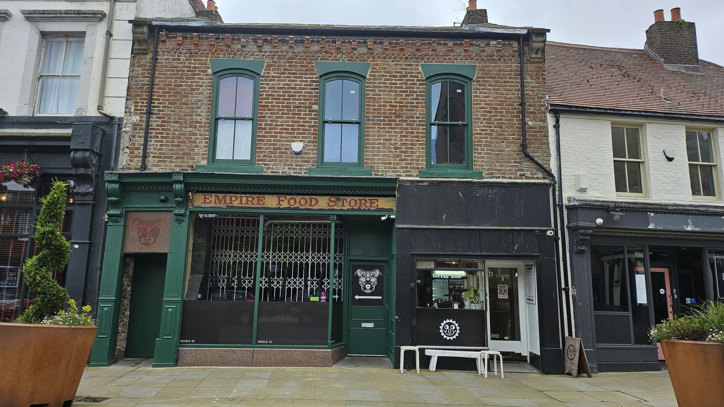 Street view of a historic brick building with green storefront and three upper windows, labeled 'Empire Food Store,' with neighboring black and white buildings and potted plants on the sidewalk.