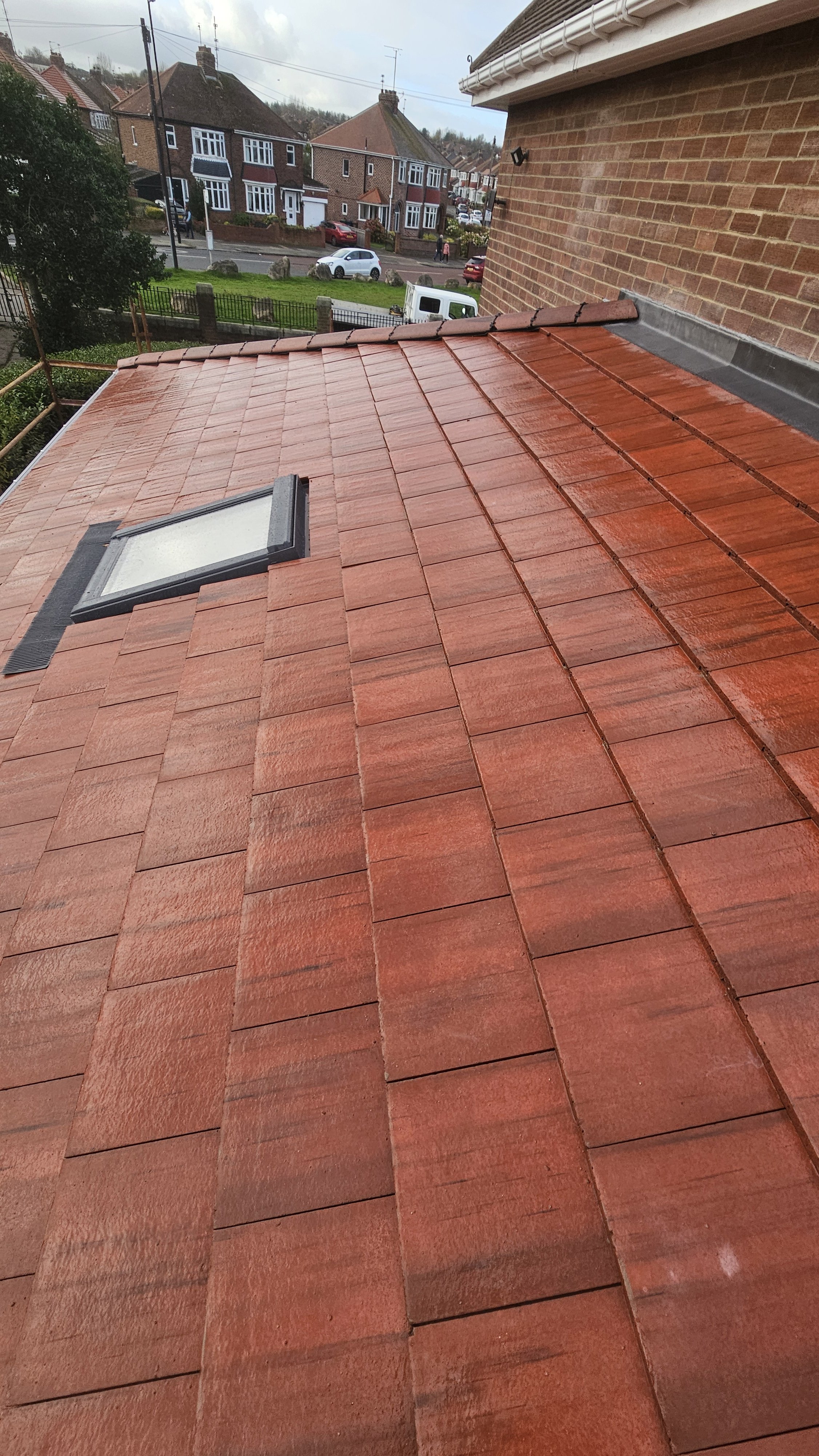 Red tiled roof with a skylight window, rain droplets visible, in a residential neighborhood with houses, cars, and street in the background.