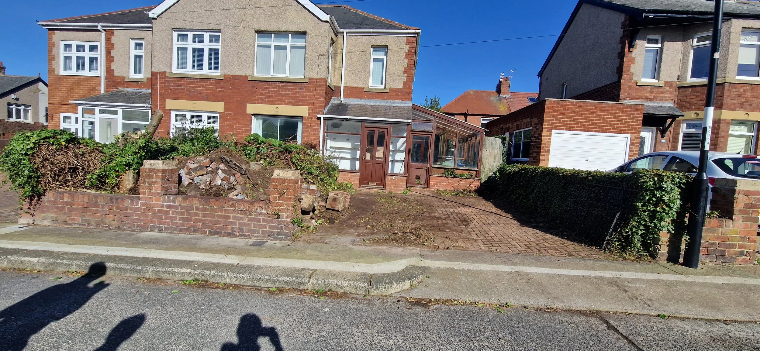 A row of brick and concrete houses with front gardens, one featuring a greenhouse, under a clear blue sky. Shadow of a person taking photo is visible on the street sidewalk.