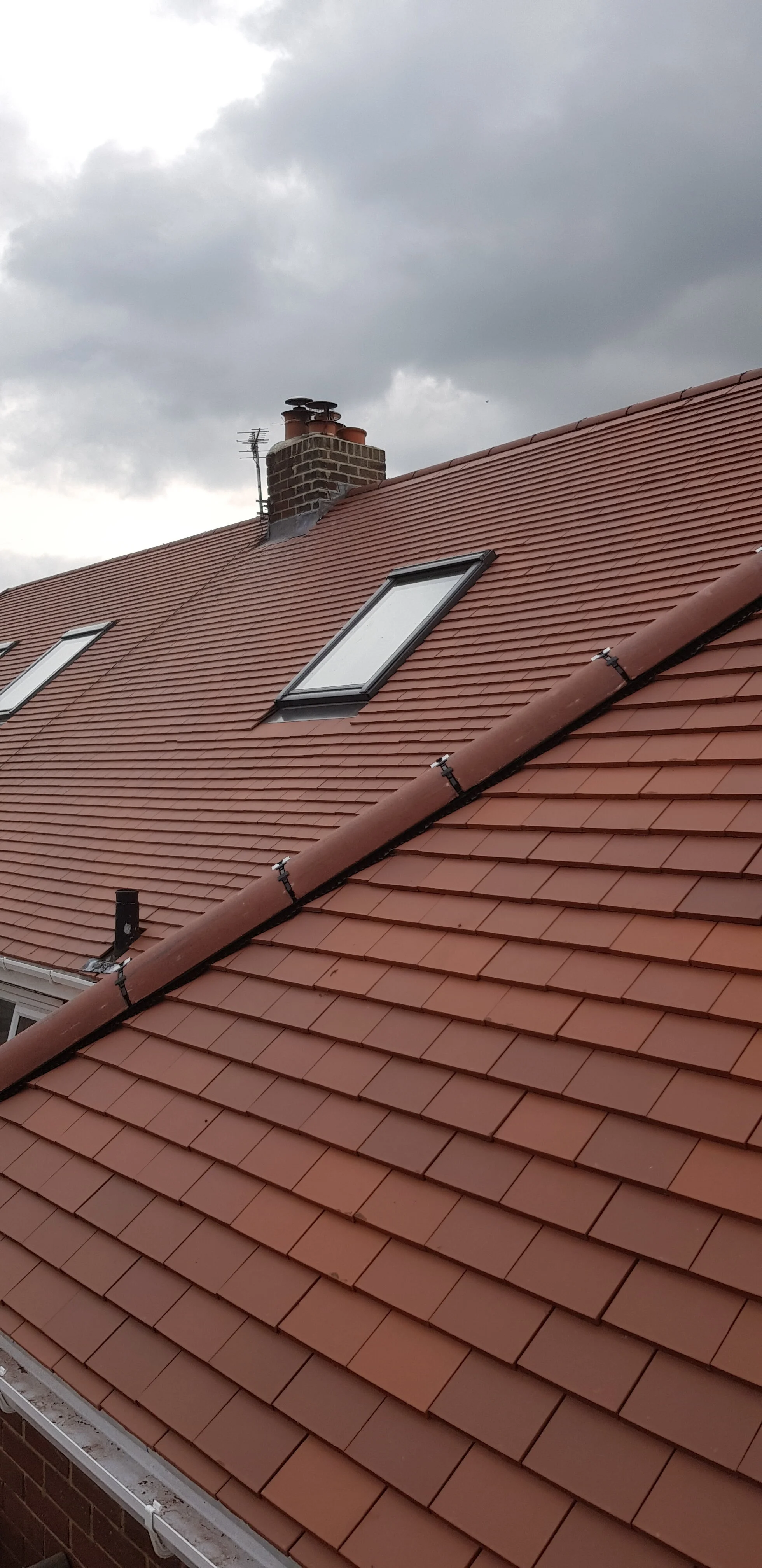 Rooftop with red clay tiles, three skylights, and a brick chimney with chimney pots, under cloudy sky.