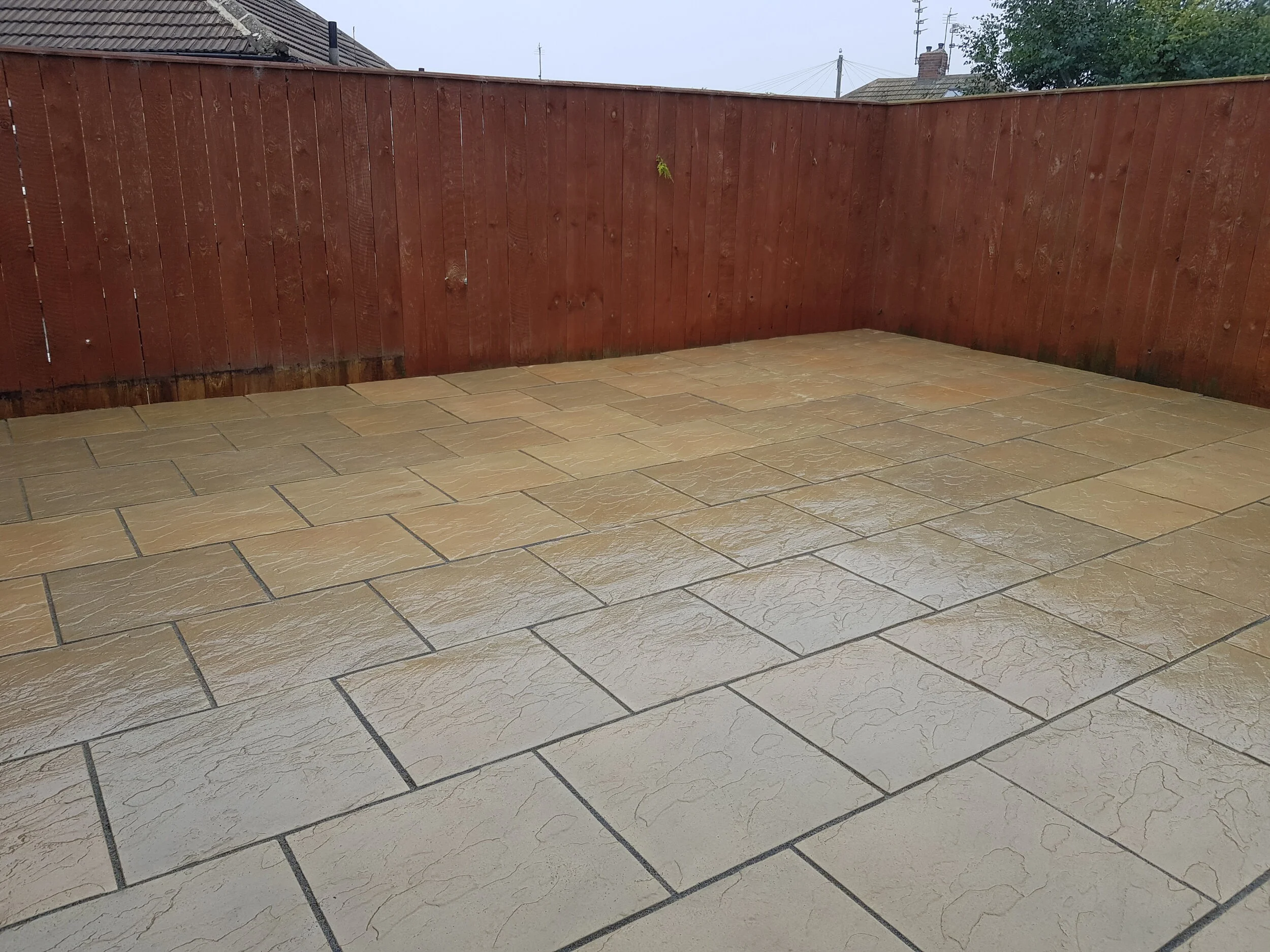 Patio with beige stone tiles and a red wooden fence on three sides, with rooftops and trees in the background.