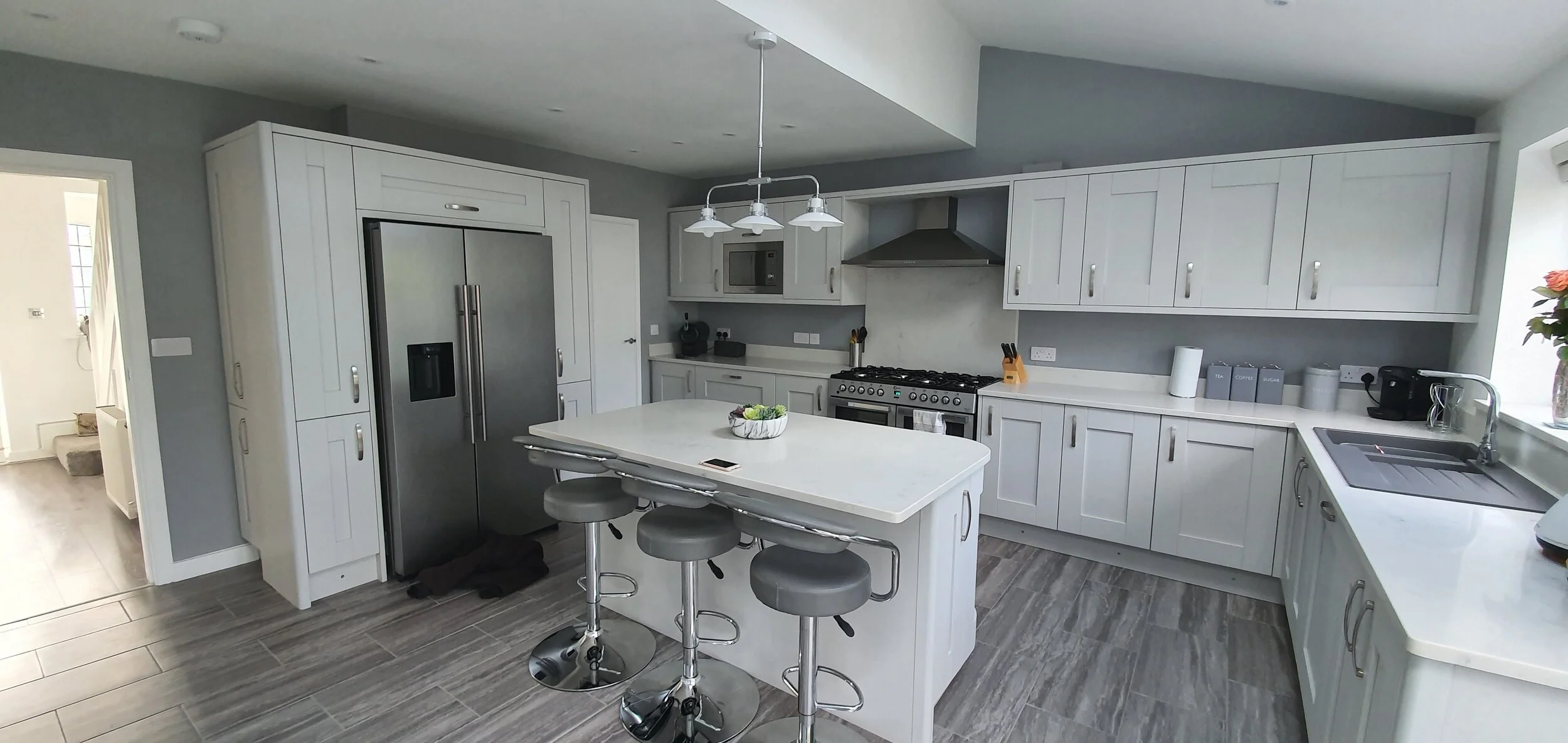 Modern kitchen with white cabinets, stainless steel refrigerator, stove, microwave, and black coffee machine. Central island with three gray bar stools, gray walls, and a window with a flower vase on the counter.