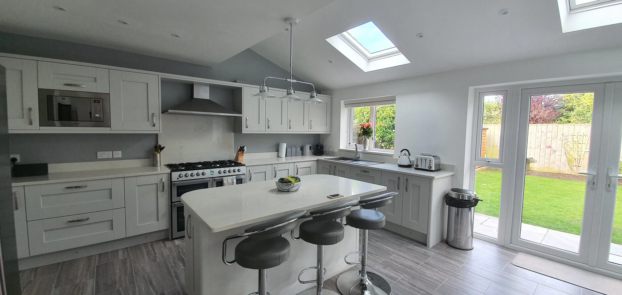 Modern kitchen with white cabinets, a central island, stainless steel appliances, and large windows and skylight providing natural light, overlooking a green backyard.