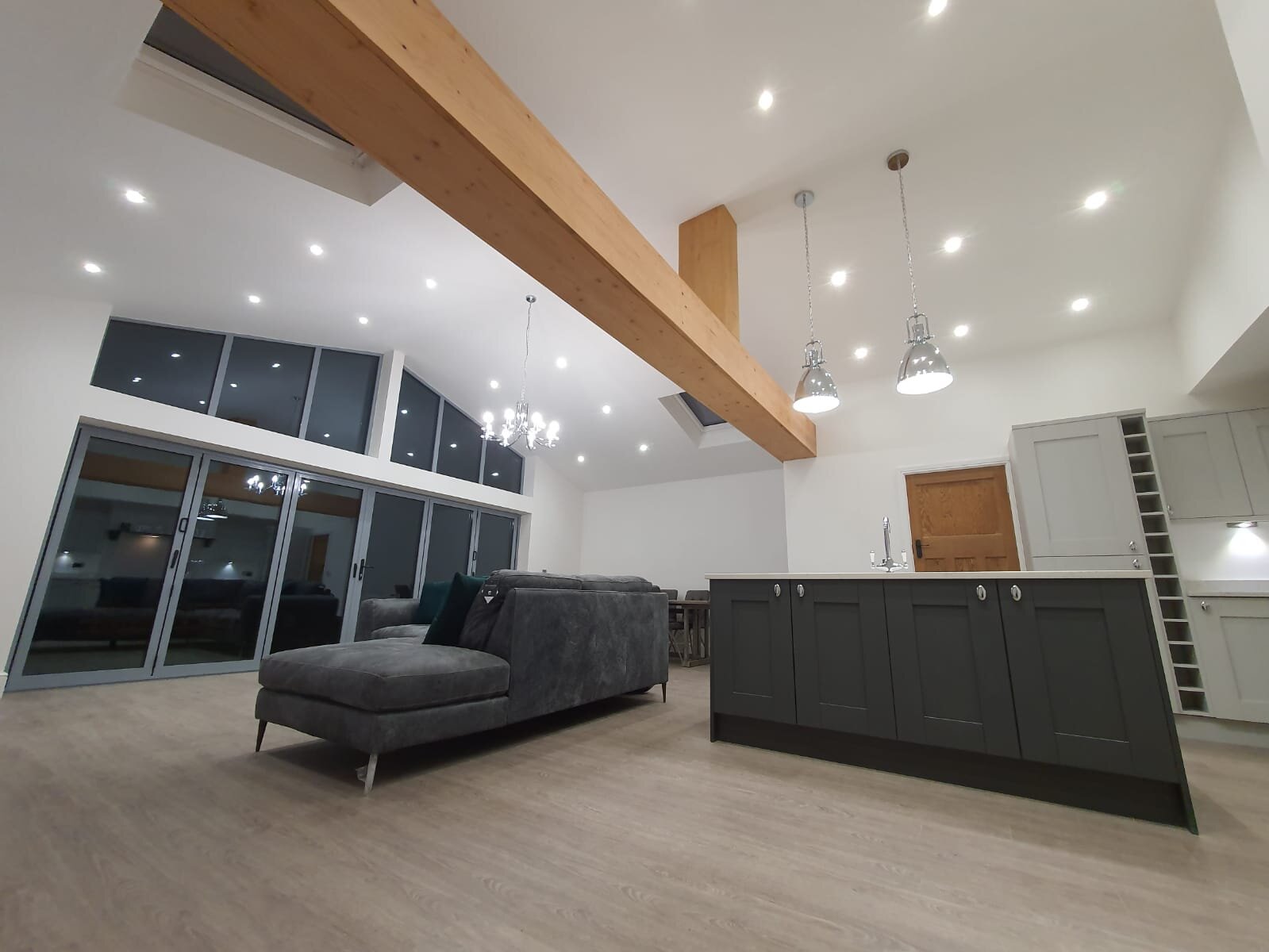 Spacious modern living room with a gray sofa, large glass sliding doors, wooden ceiling beam, and a kitchen island with pendant lighting.
